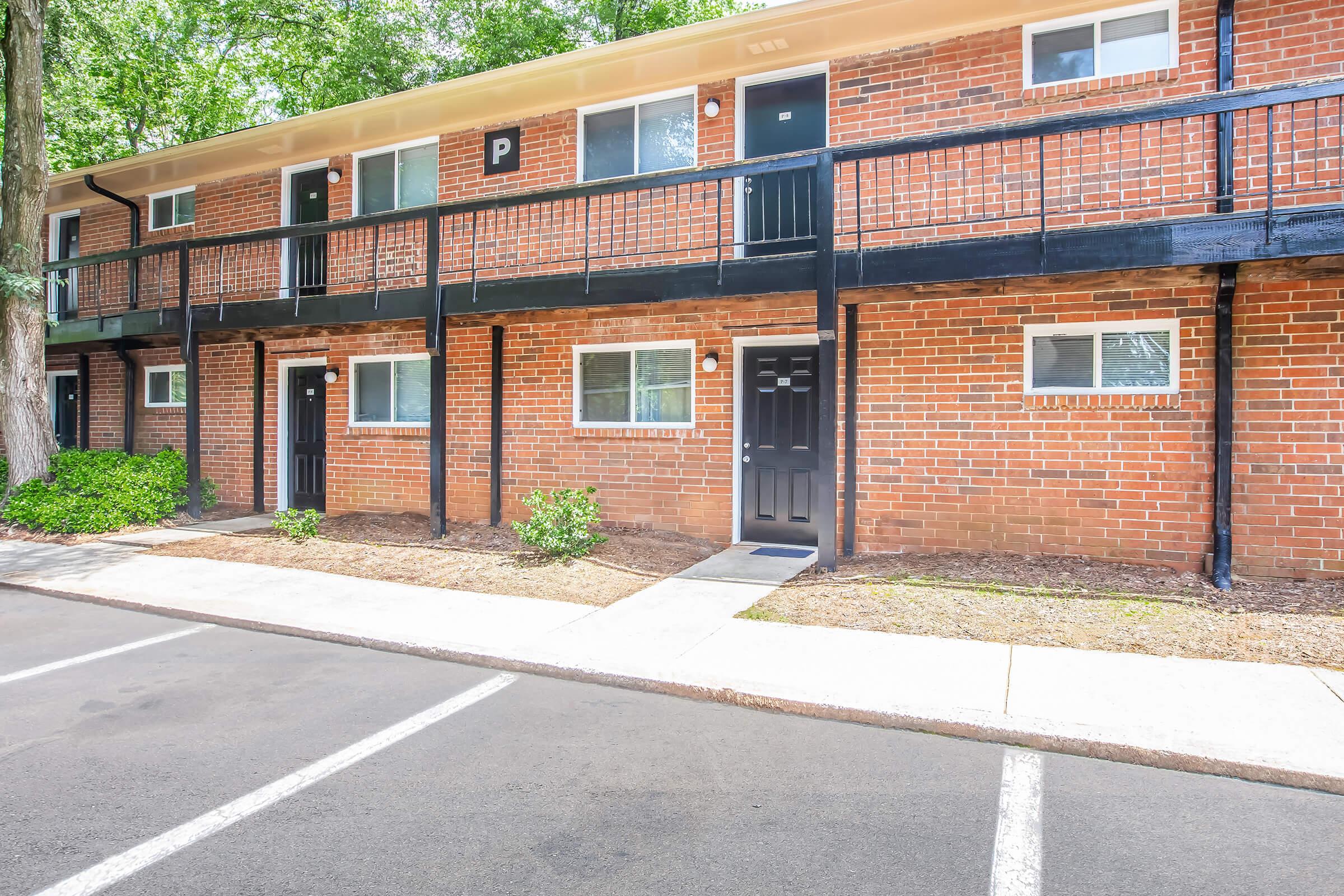 A view of a brick apartment building featuring two levels with black doors and railings. The ground is landscaped with greenery beside a walkway leading to the entrance. A parking area is visible in the foreground with empty spaces.
