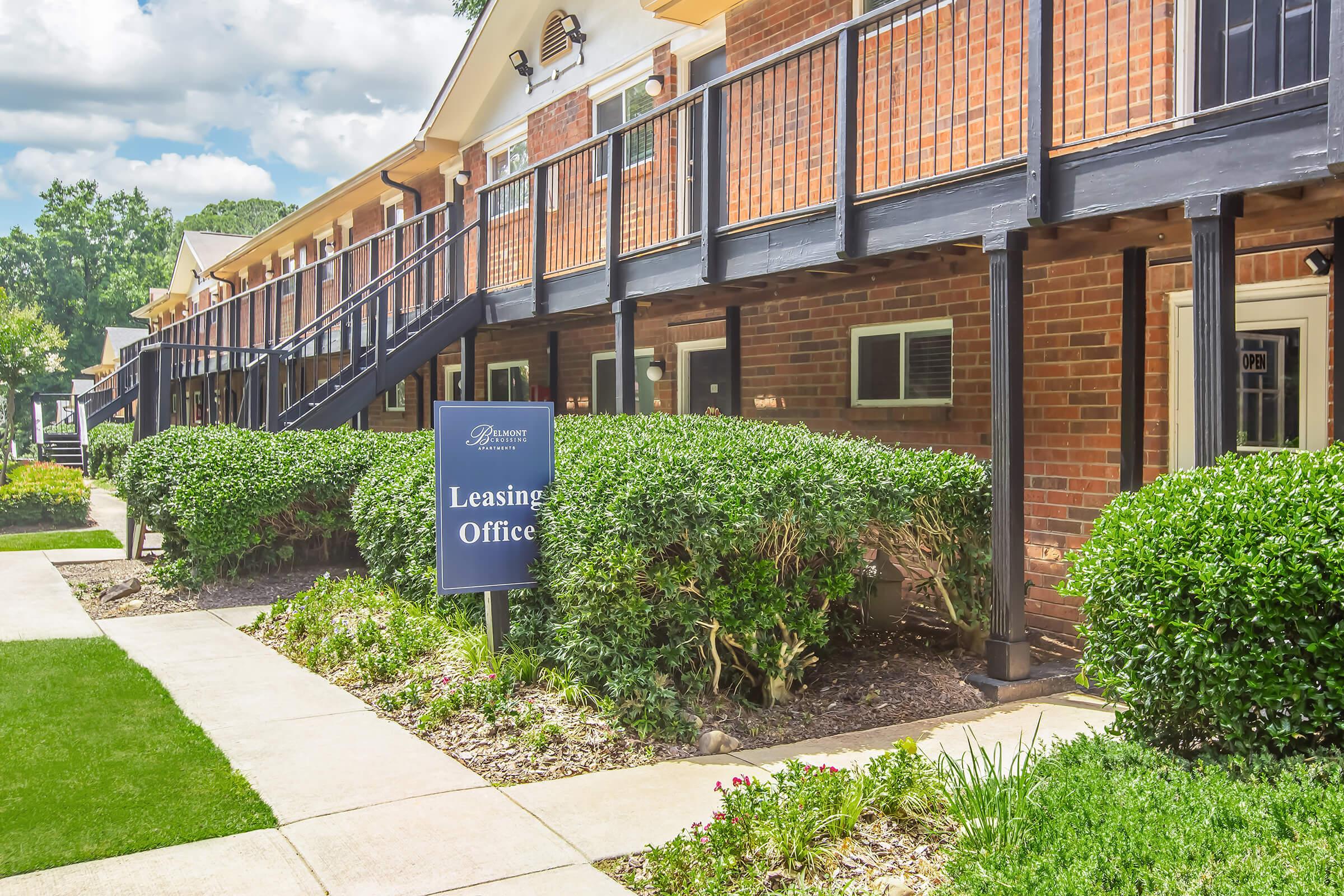 A well-maintained apartment complex with a leasing office sign in front. The path is lined with manicured shrubs, and the building features brick exteriors and balconies. Clear blue skies and fluffy clouds are visible, adding to the inviting atmosphere.