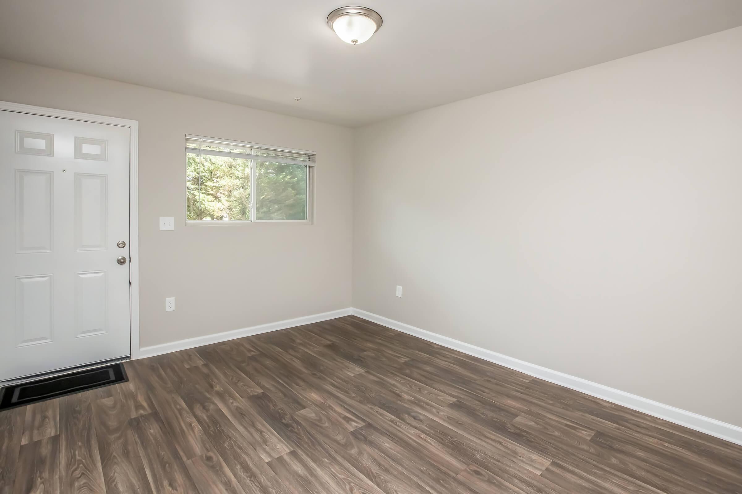 Empty room with light wood laminate flooring, a window providing natural light, and a front door. The walls are painted in a neutral color. No furniture or decorations are present, creating a clean and minimalist appearance.