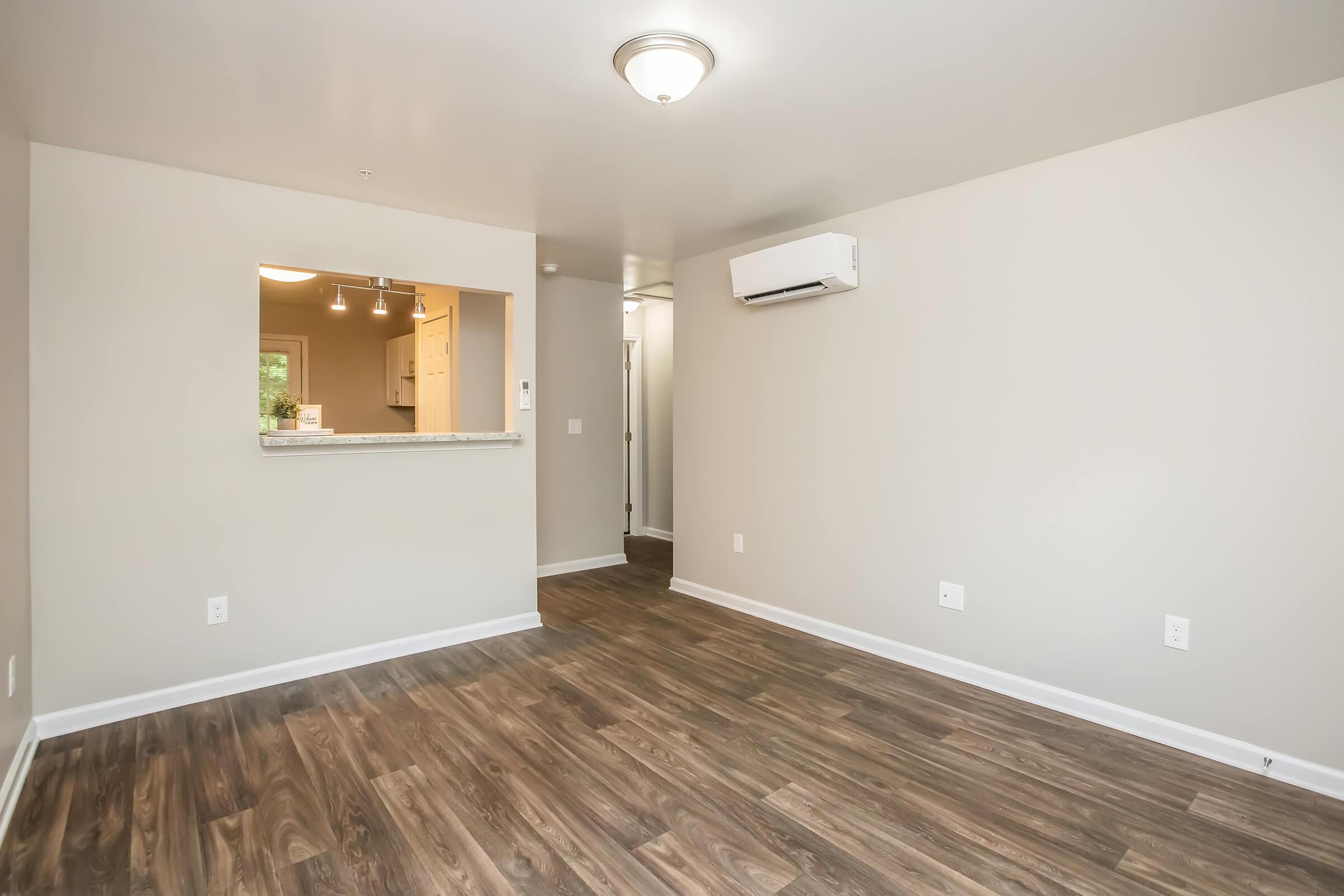 A spacious, empty living room with light-colored walls and dark wood-style flooring. There is an air conditioning unit on one wall, and a doorway leading to another room. An open archway leads to a kitchen area with bright lighting.