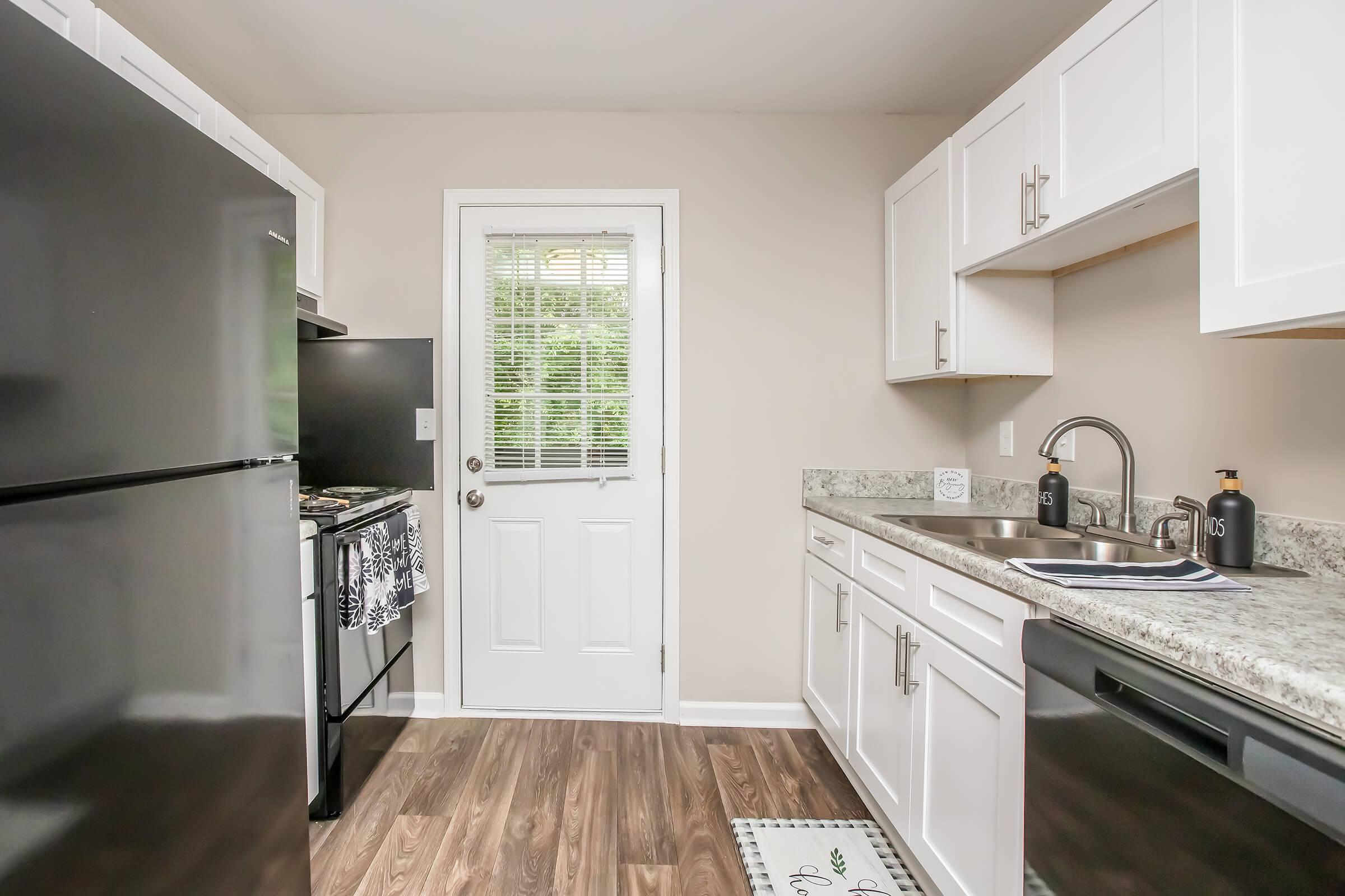 Modern kitchen featuring a black refrigerator and stove, white cabinetry, and a granite countertop. There's a sink with stylish fixtures and a small door leading outside. Natural light comes through a window, highlighting the wooden floor and overall design.
