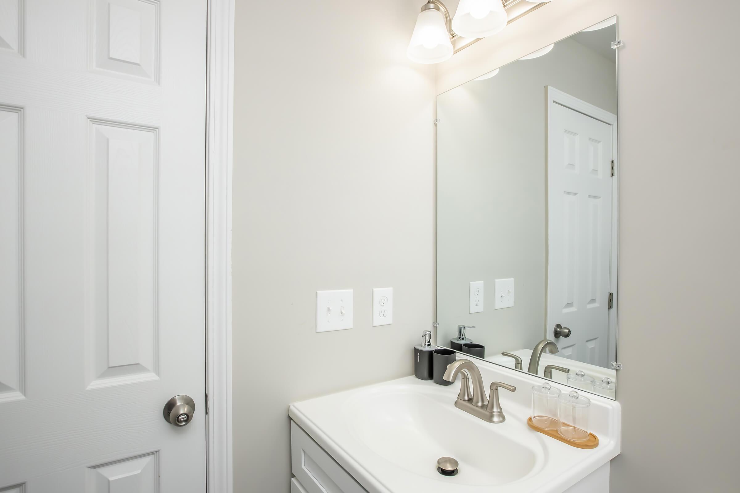 A clean bathroom featuring a white vanity with a sink, polished faucet, and two glasses on a wooden tray. A large mirror above the sink reflects the bright light from three fixtures. A white door is visible to the left, contributing to a modern, minimalistic aesthetic.