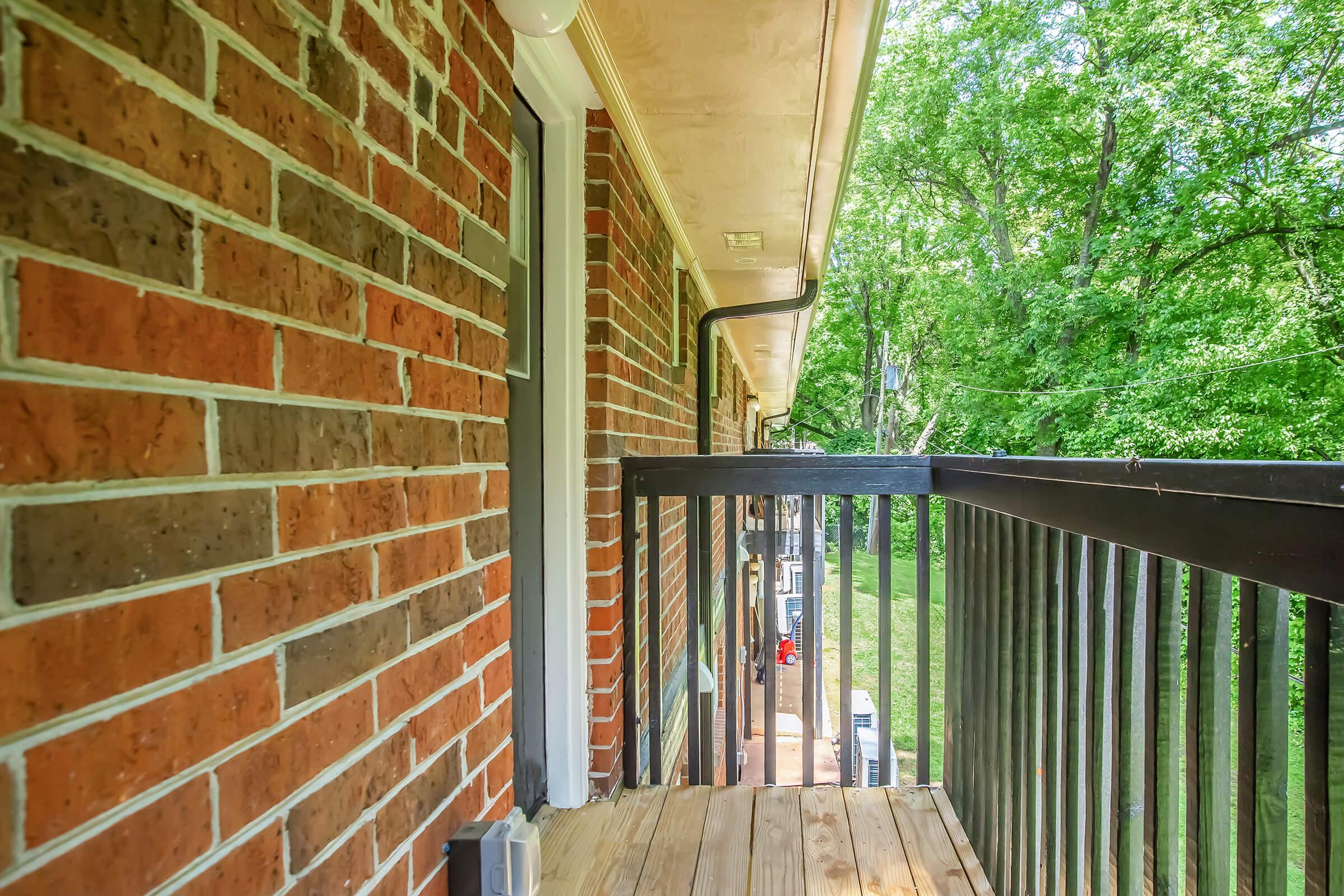 A view of a balcony with wooden flooring, surrounded by a brick wall, overlooking a lush green garden. The balcony features black railings and a door leading indoors. Sunlight filters through the trees in the background, creating a serene and inviting atmosphere.