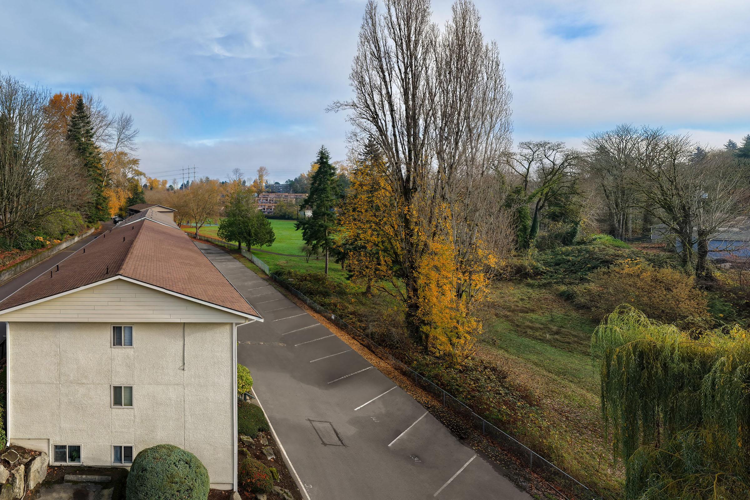View from an elevated position showing a residential building to the left, an empty parking lot, and a green field surrounded by trees in autumn colors. The sky is clear with soft clouds, and the landscape is serene, blending natural and urban elements.