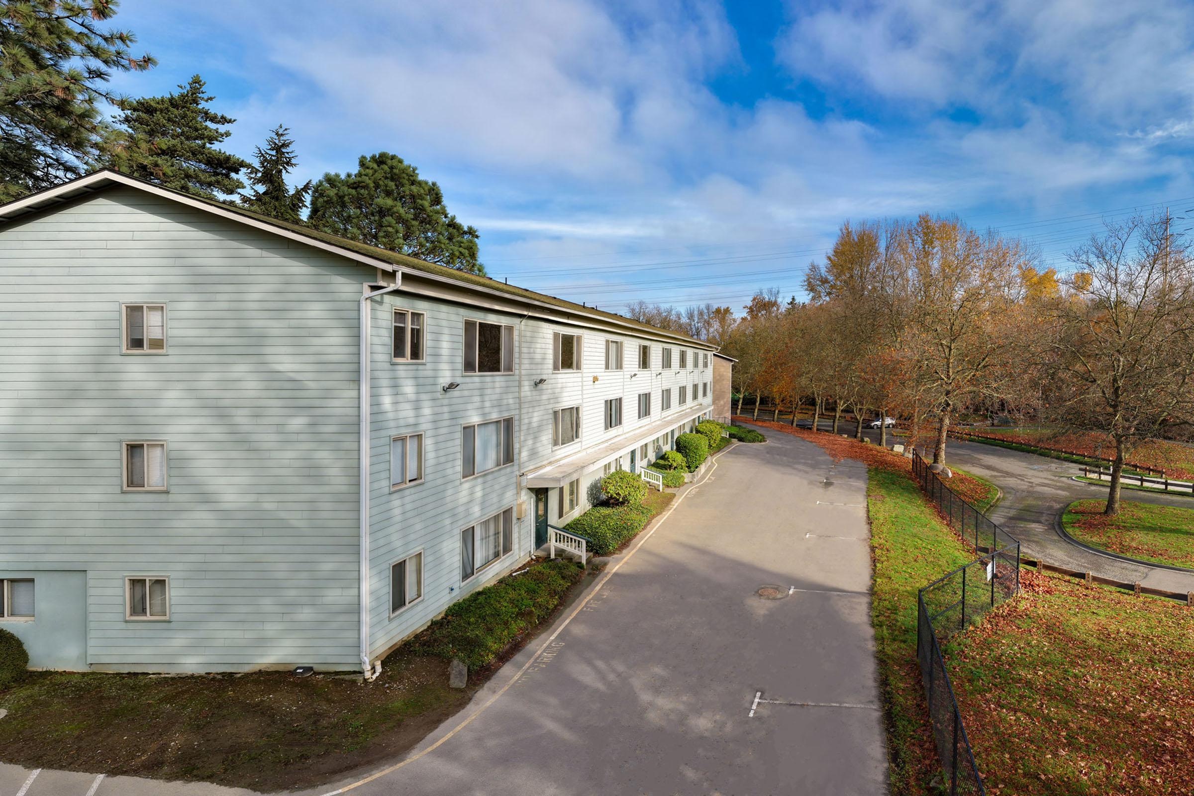 A light blue three-story building surrounded by trees, with a paved path leading to a parking area. The scene features a clear sky and hints of autumn foliage along the pathway.