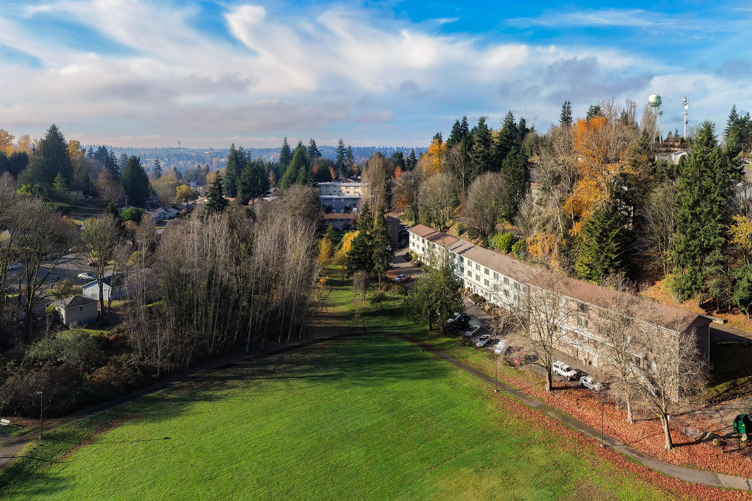 Aerial view of a residential area surrounded by trees, featuring low-rise buildings and parked cars. The foreground displays a spacious green lawn, while the background shows neighborhoods, hills, and a clear blue sky with scattered clouds. Autumn foliage adds warmth to the landscape.