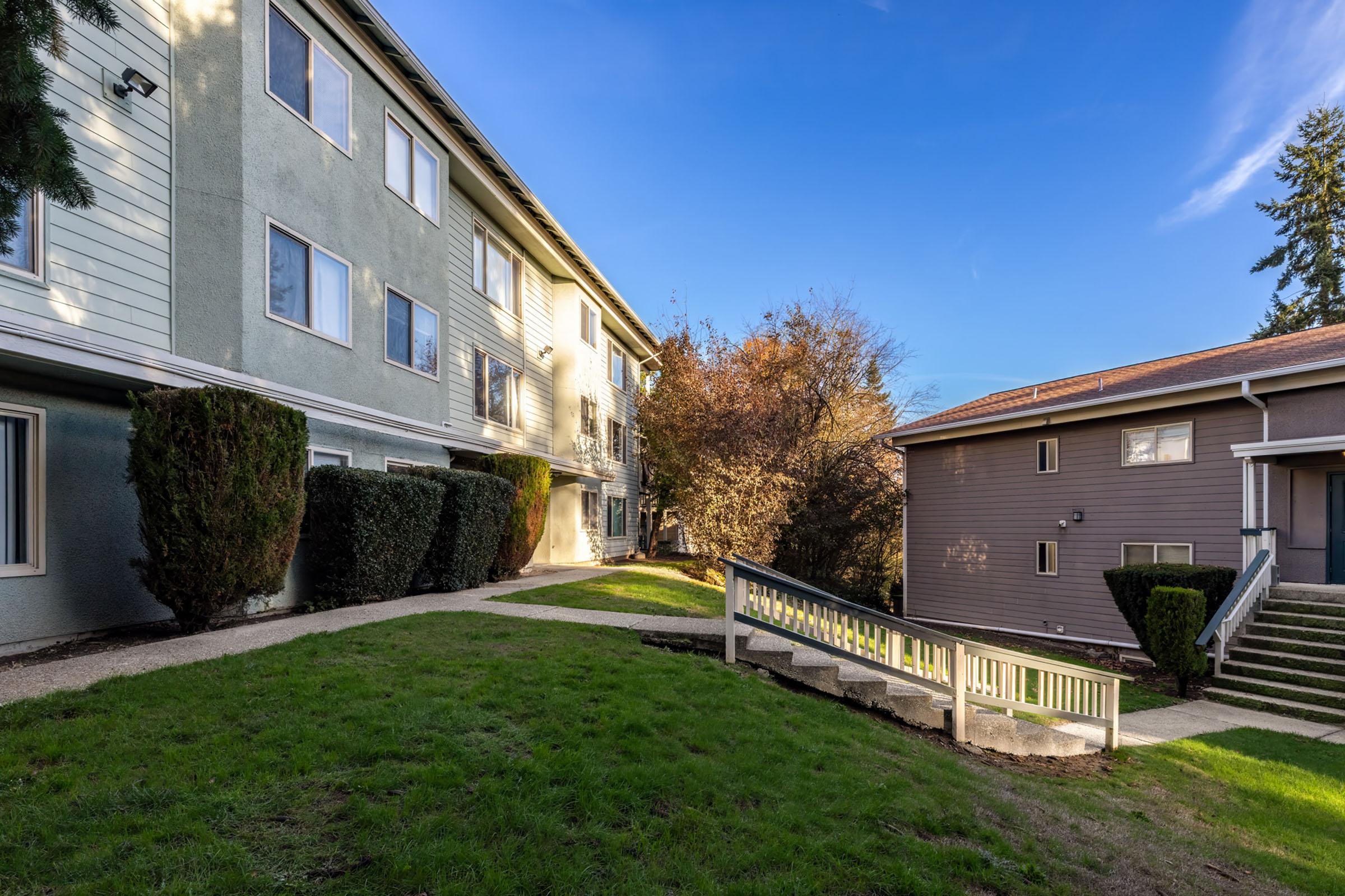 A landscaped area featuring two multi-story buildings with different architectural styles. One building has green and white siding, and the other is gray. There are neatly trimmed bushes and a grassy path leading to a set of stairs. The sky is clear and blue, adding a pleasant atmosphere to the scene.