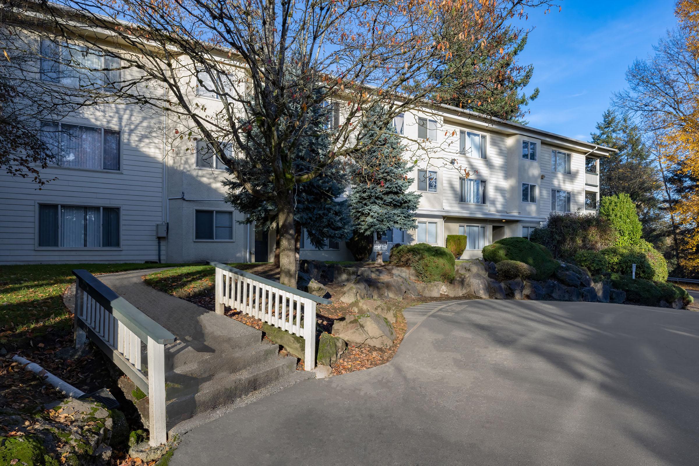 A multi-story residential building surrounded by trees and landscaped shrubs. A pathway with a ramp leads to the entrance, which has several windows reflecting the blue sky. The scene is set in autumn, with colorful foliage and a clear, sunny sky.