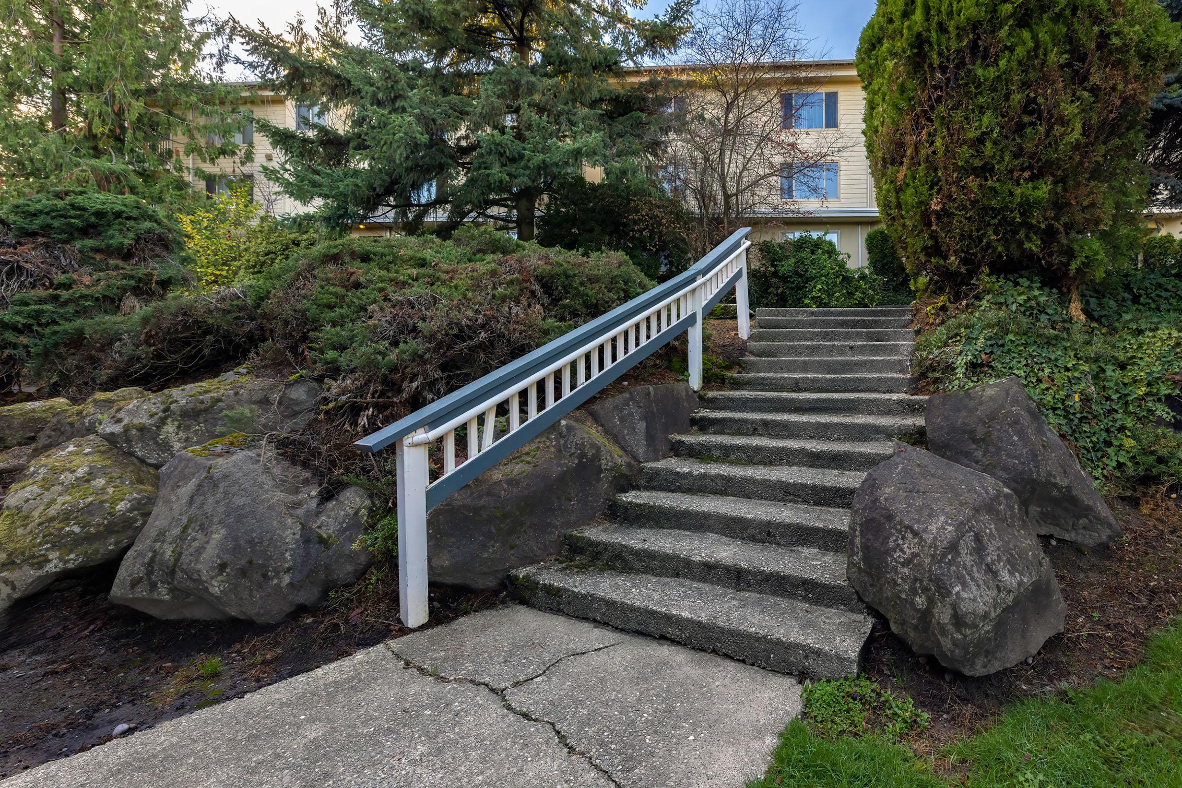 A stone pathway leading up a set of stairs, flanked by large rocks and lush greenery. A white railing runs along the stairs, which ascend toward a building partially visible in the background. The scene is tranquil, with vibrant plants and a clear sky.