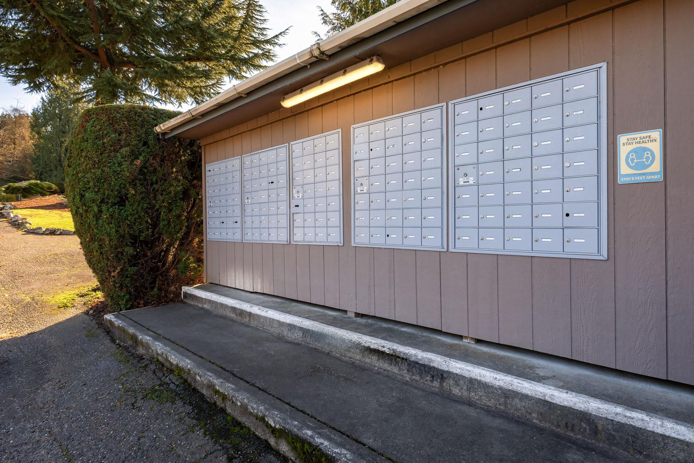 A postal mailbox unit mounted on an exterior wall with multiple individual mail slots. The building is surrounded by greenery, and there is a light fixture overhead. The concrete slab in front provides access to the mailboxes, set in a residential area.