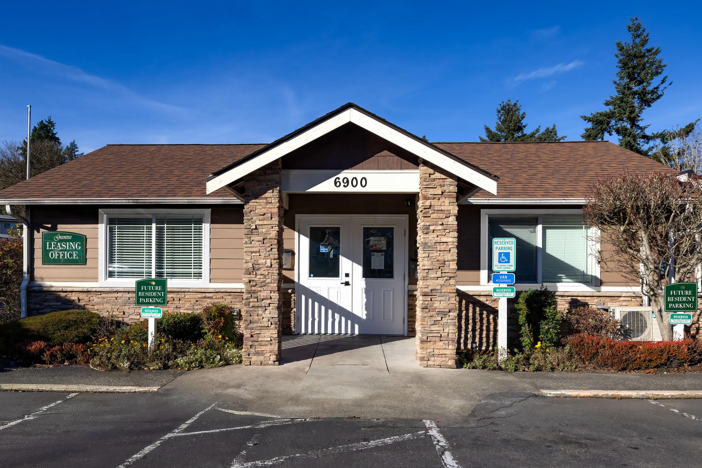 A single-story leasing office building with a brown shingle roof and stone accents. The front has a sign displaying "Leasing Office" and the number "6900." There are two accessible parking signs and landscaped greenery around the entrance, with clear blue skies in the background.