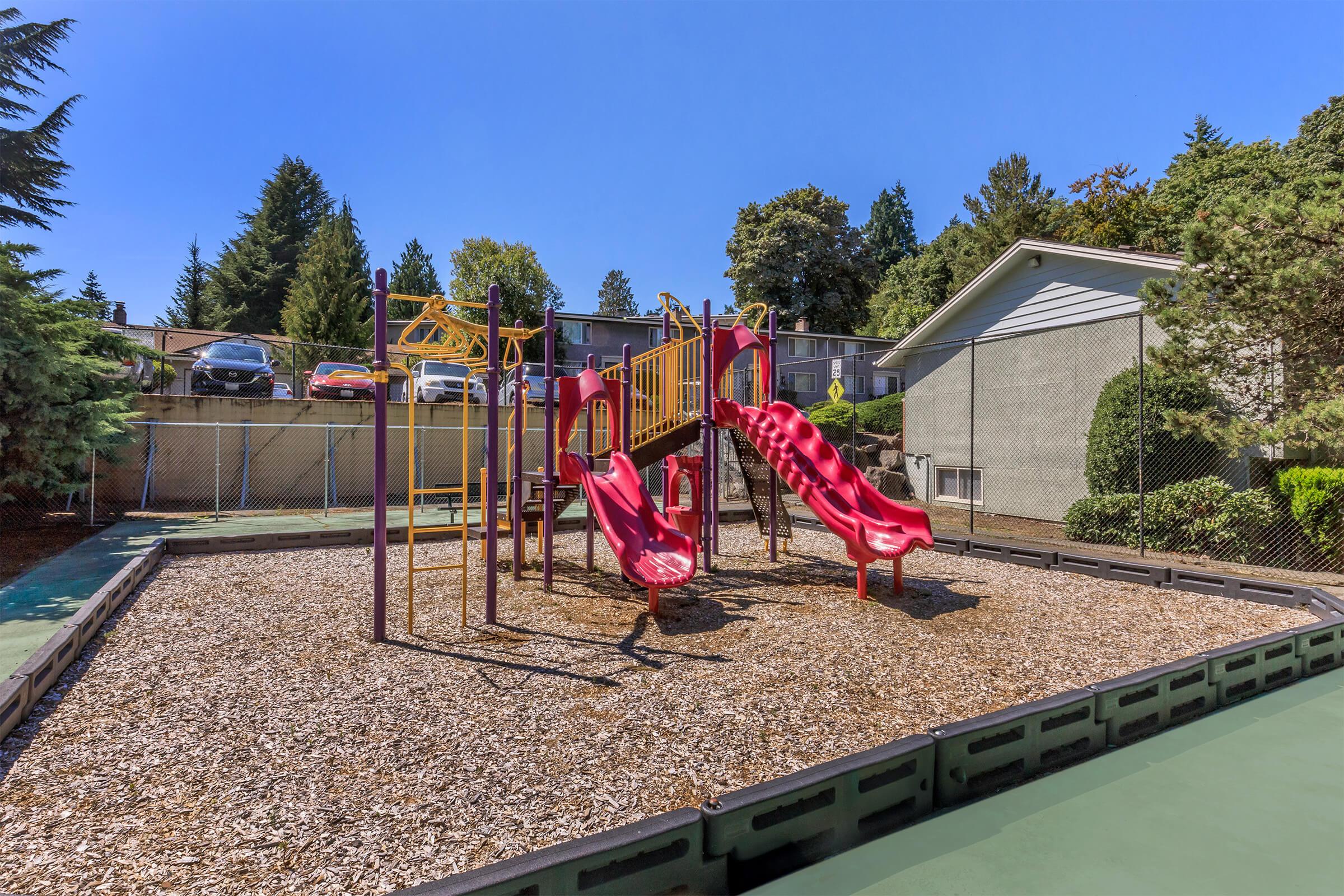 Playground area featuring two red slides, climbing structures, and soft ground cover made of wood chips. The background shows a fenced area with trees and a residential building nearby. Clear blue sky overhead and a well-maintained environment suggest a welcoming space for children.