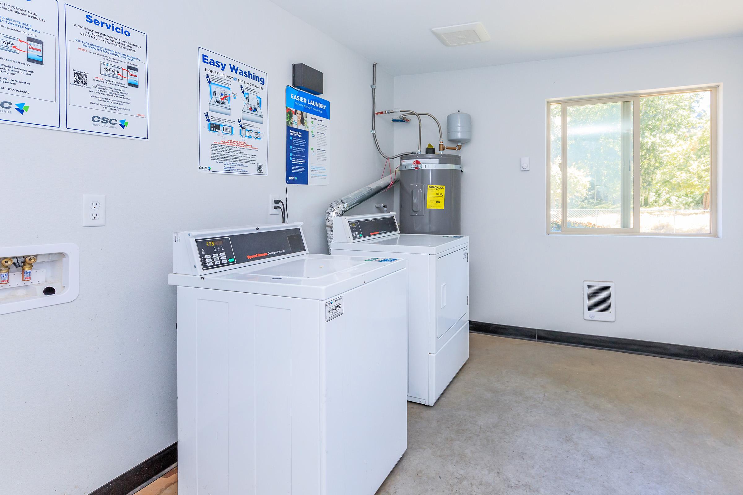 A clean, well-lit laundry room featuring two washing machines and a dryer. There are informational posters on the wall, a water heater visible, and a window providing natural light. The floor is concrete, and the overall space is organized and functional.