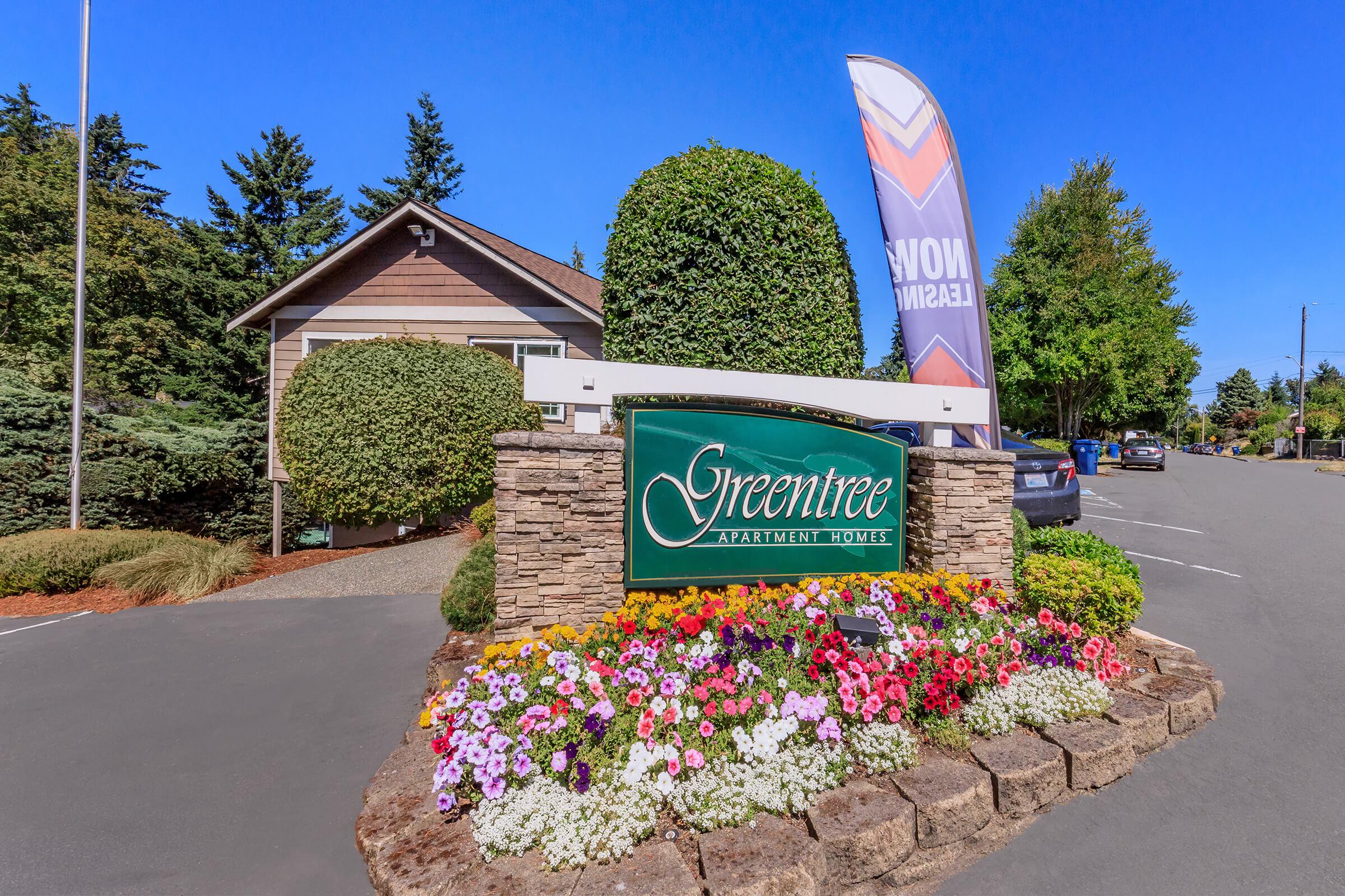 Sign for Greentree Apartment Homes surrounded by colorful flowers and neatly trimmed hedges. A large banner reads "Now Leasing" next to the entrance. In the background, there are trees and parked cars on a clear day.