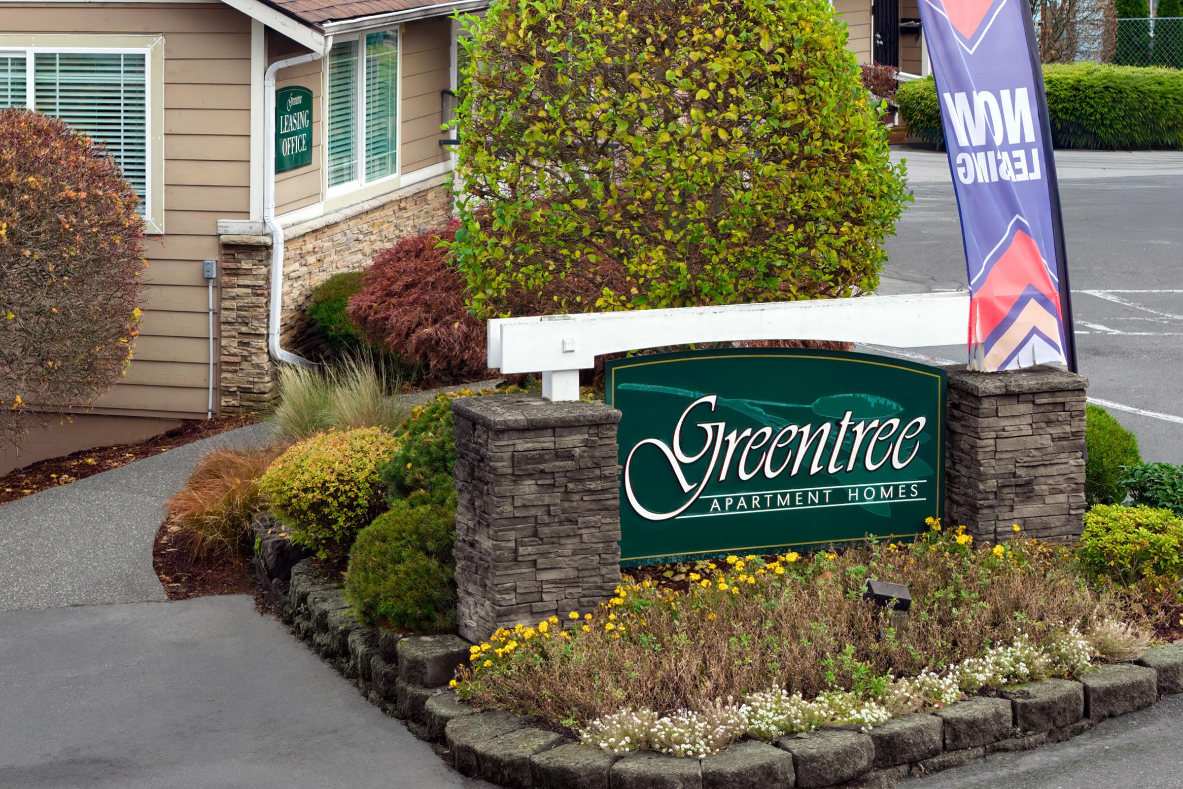 Sign for Greentree Apartment Homes featuring lush landscaping and a colorful flag indicating hiring. The entrance is well-maintained with flowers and greenery surrounding the stone base of the sign, set against a backdrop of residential buildings.