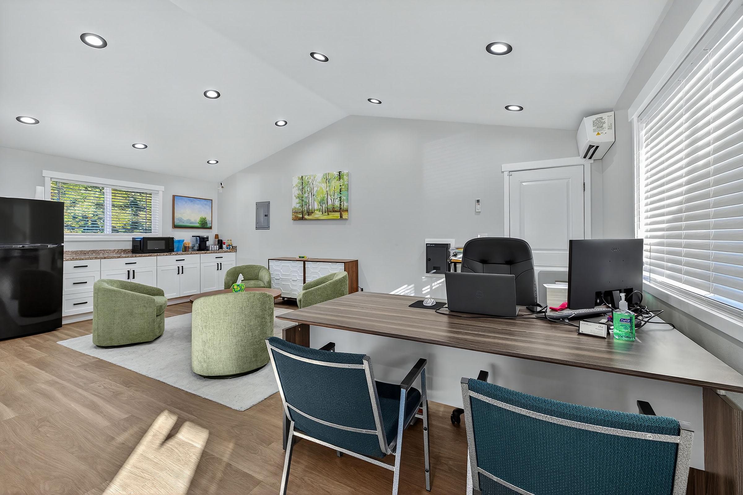 A bright, modern office space featuring a wooden desk with a computer and a chair. There are two green armchairs and a white rug on the floor. The kitchen area includes a black refrigerator and a cabinet. Large windows provide ample natural light, and a landscape painting is hung on the wall.