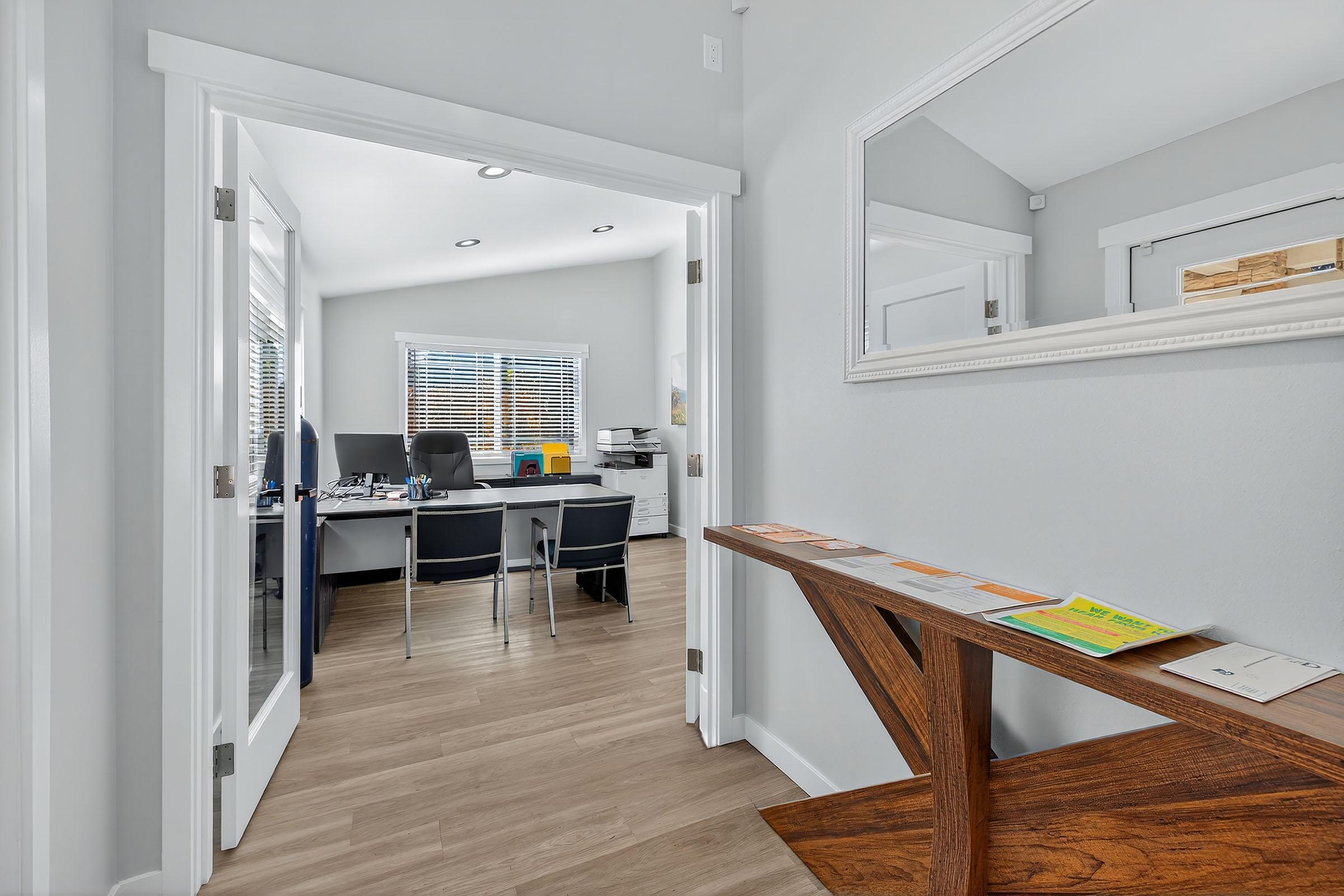 A spacious hallway leads to a modern office with a desk and ergonomic chair, surrounded by windows providing natural light. A stylish wooden console table with colorful brochures is positioned in the foreground, enhancing the welcoming atmosphere. The walls are painted in soothing gray tones.