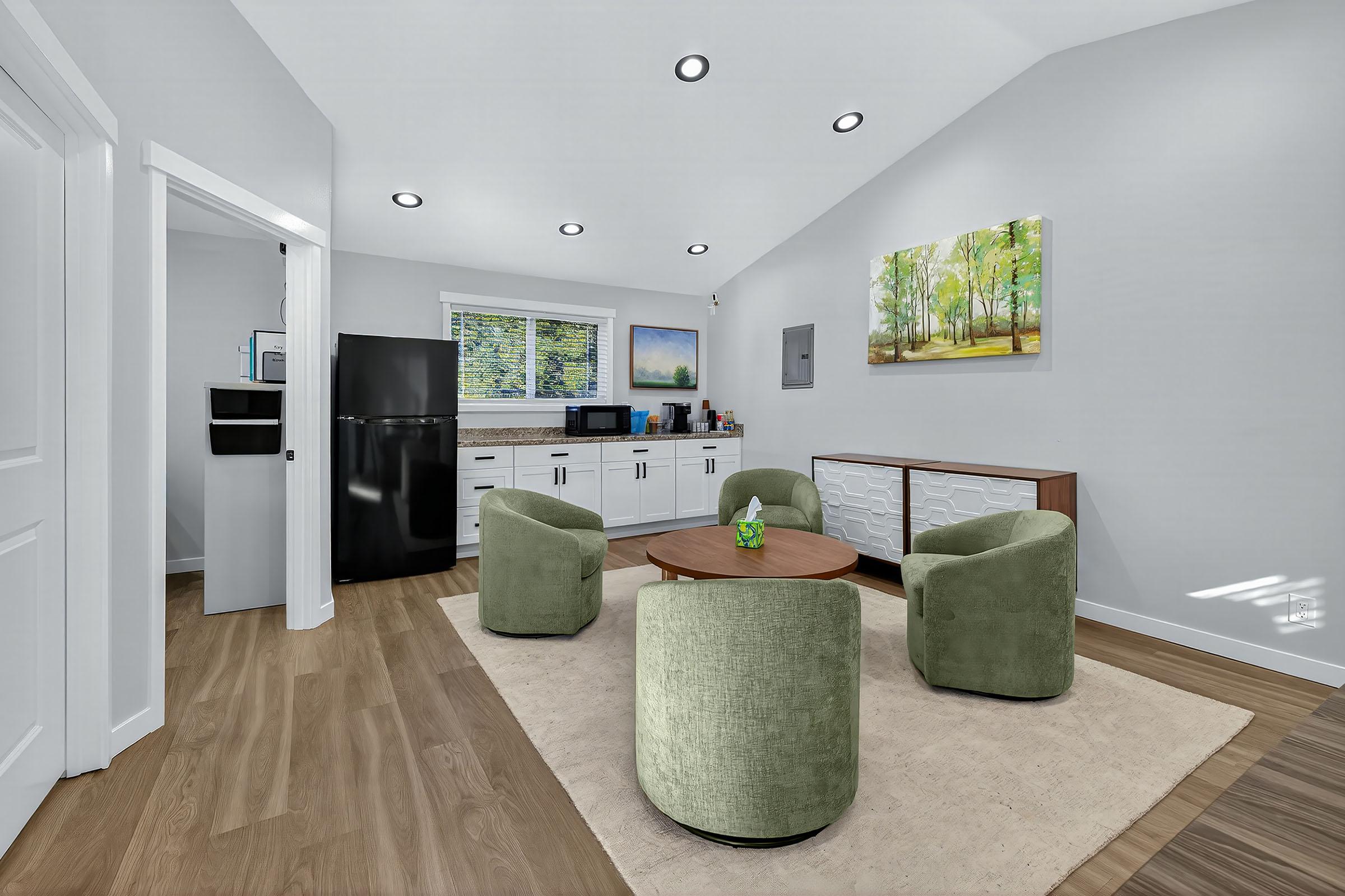 Modern kitchen and living area featuring a black refrigerator, microwave, and white cabinets. There are two green upholstered chairs and a wooden coffee table on a light rug. A painting of a forest scene hangs on the wall, and sunlight streams through a window. The floor is wood, creating a cozy atmosphere.