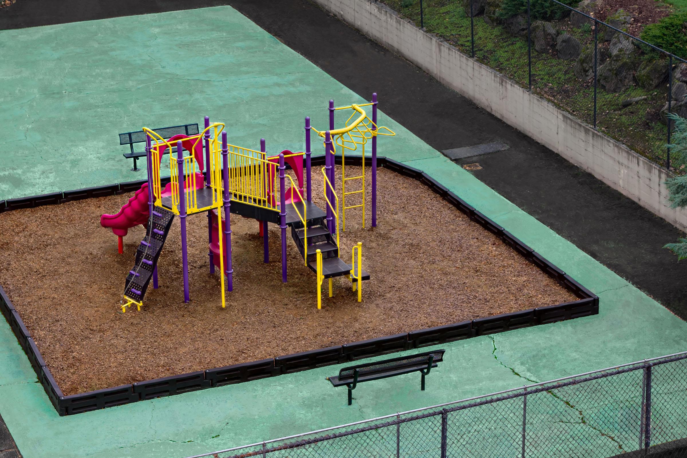 Colorful playground equipment with slides and climbing structures, surrounded by a gravel area. The playground is set on a green surface, with a bench nearby and a fenced perimeter, seen from an elevated angle.
