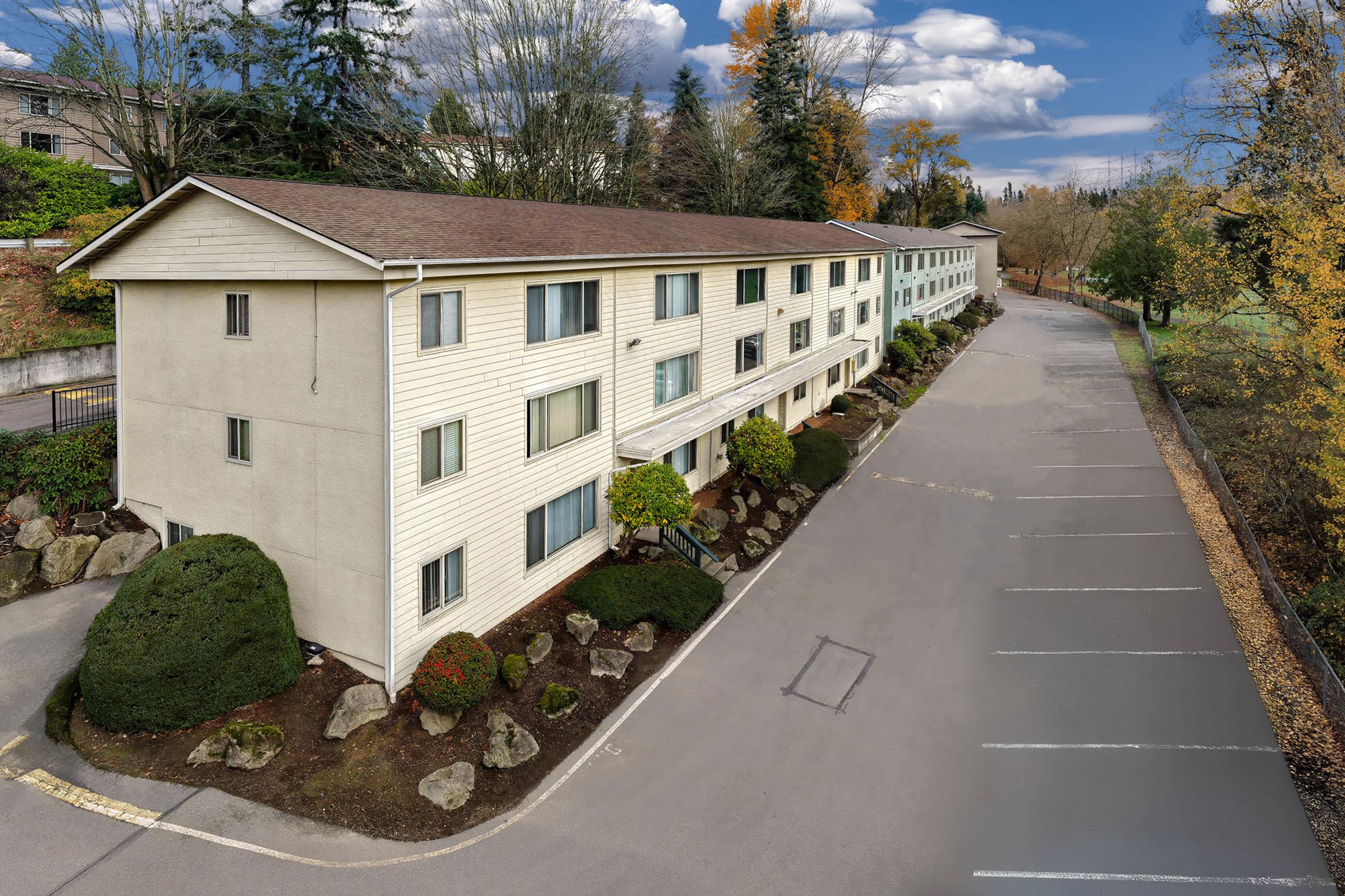 Aerial view of a multi-story residential building with a light-colored exterior, surrounded by neatly landscaped bushes and rocks. A paved parking area is visible in front of the building. Trees with autumn foliage are present in the background under a partly cloudy sky.