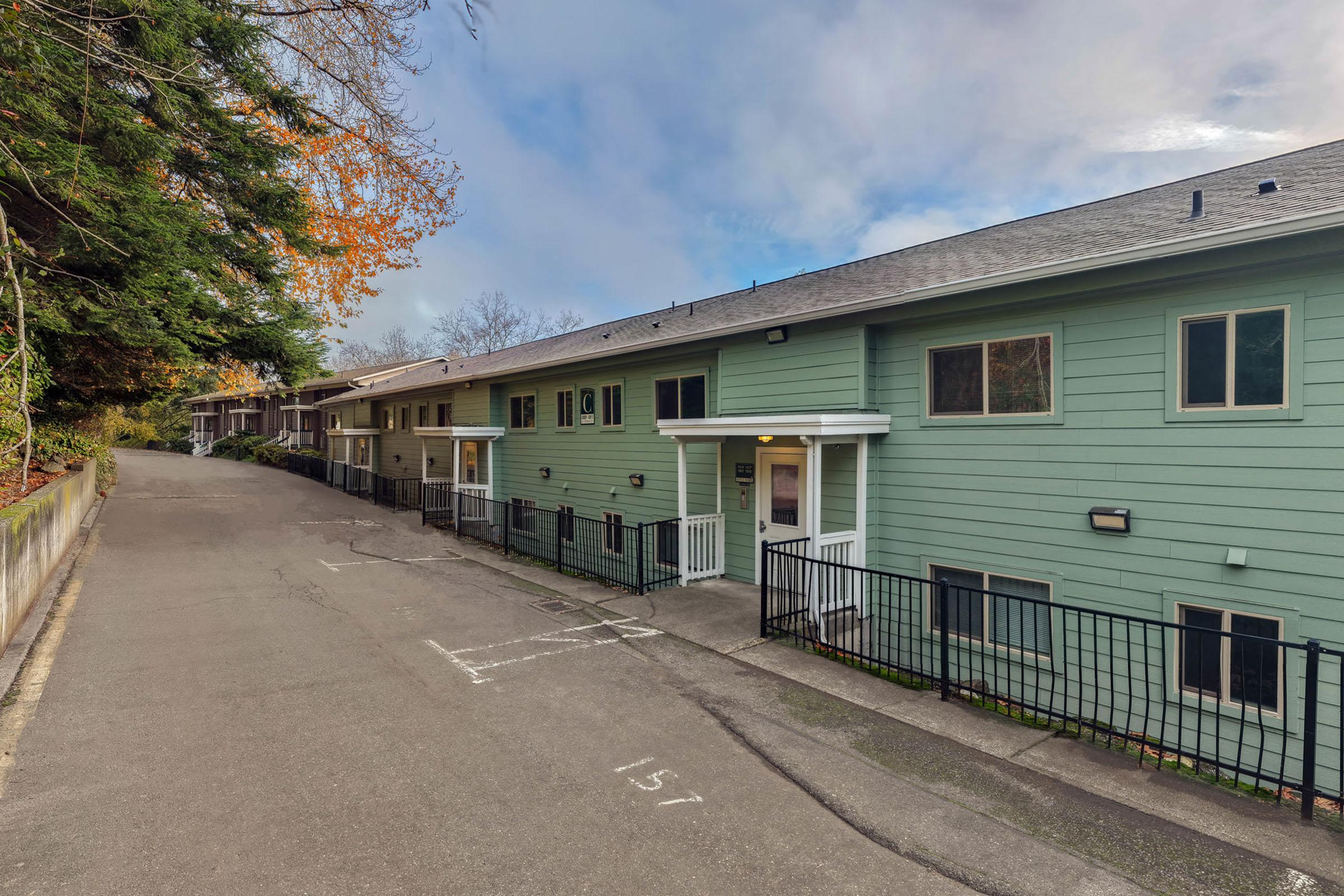 Exterior view of a row of residential buildings along a paved pathway. The buildings are painted in light green, with balconies and a white entrance. A tree line borders the area, and parking spaces are visible in front of the units. The sky is partly cloudy, creating a serene atmosphere.