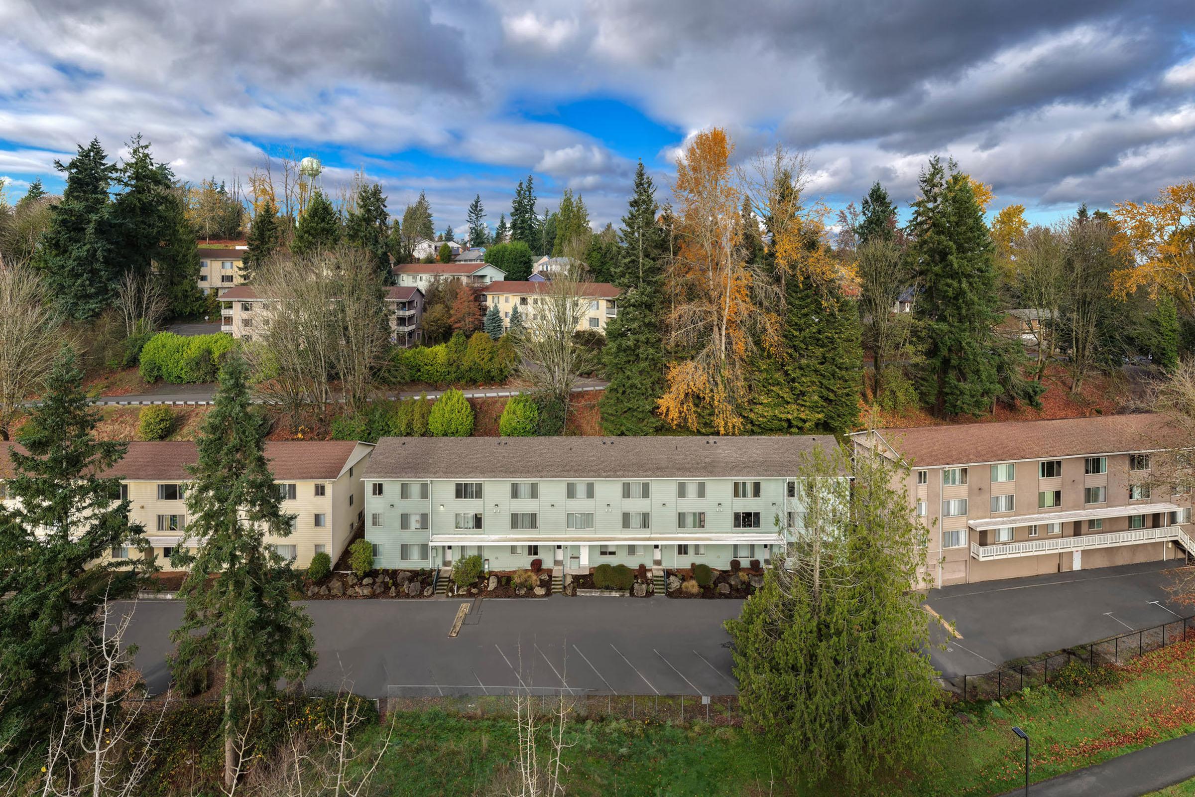 Aerial view of a multi-story residential complex surrounded by trees and colorful autumn foliage. The buildings feature a combination of light and dark exteriors, with some parking spaces visible in the foreground. A cloudy sky with hints of blue can be seen above the scene.