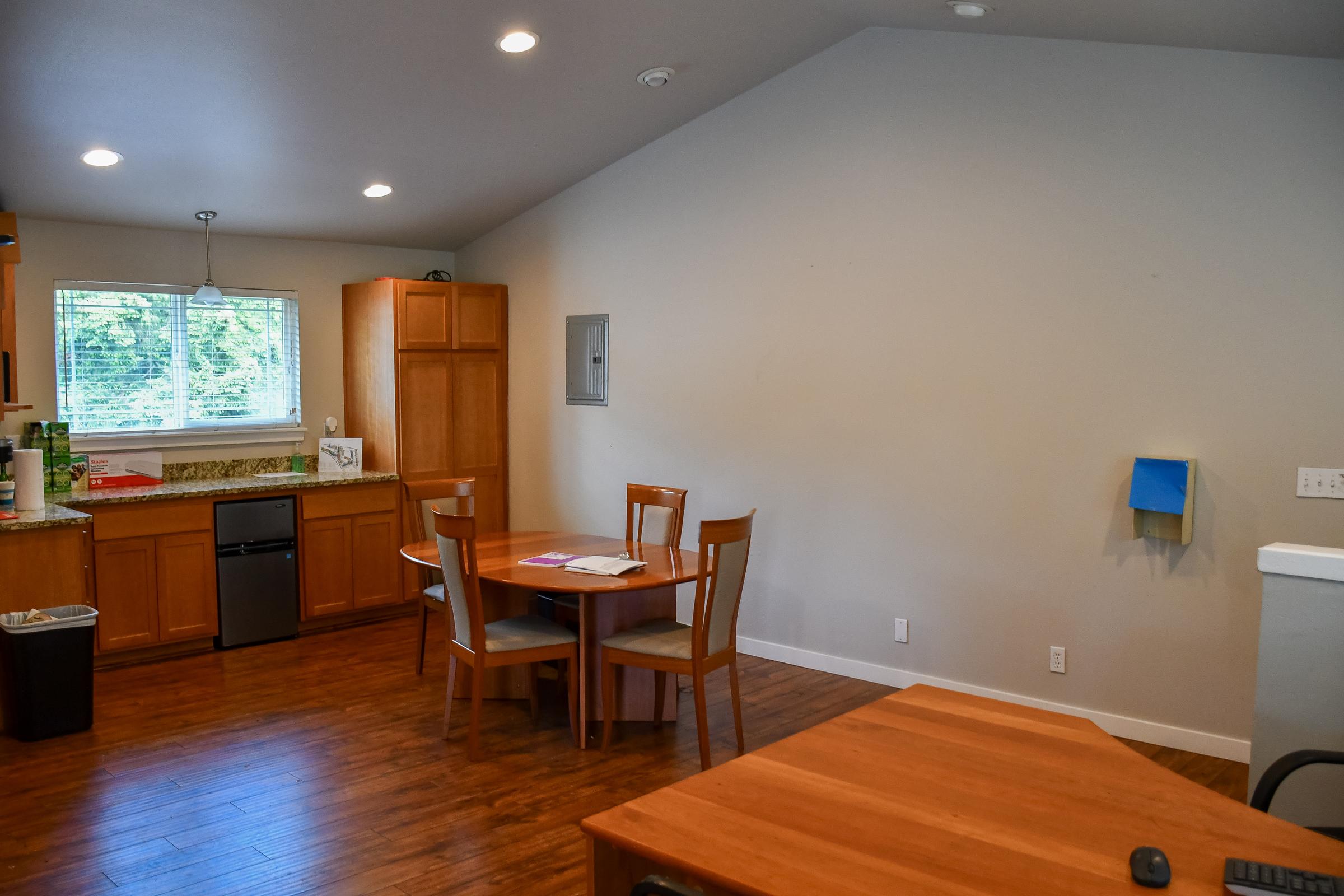 A modern kitchen and dining area featuring wooden cabinets, a round dining table with four chairs, a stainless steel dishwasher, and large windows letting in natural light. The space has hardwood floors and minimal decor, providing a clean and open atmosphere.
