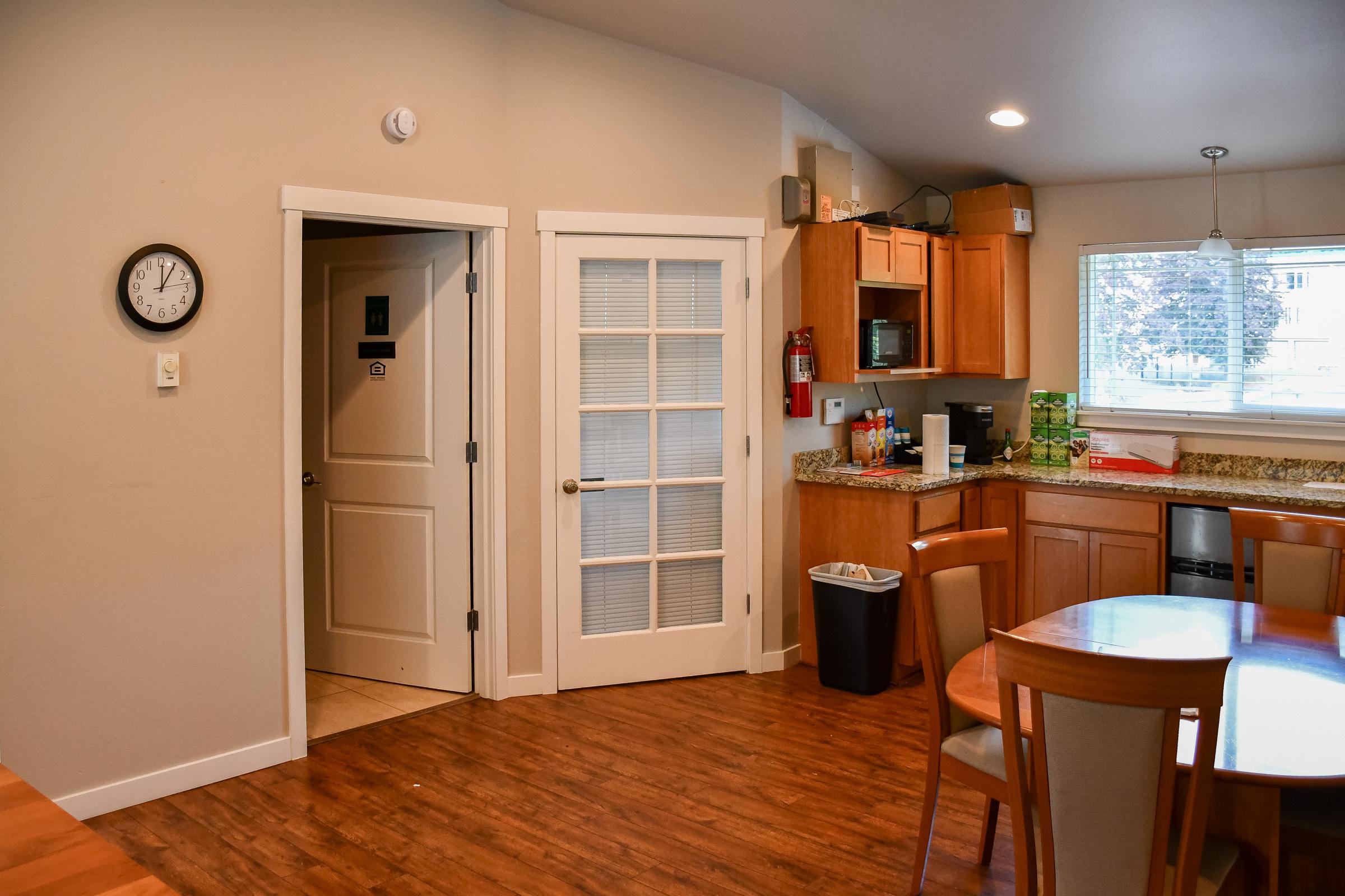 A cozy kitchen and dining area with wooden floors, featuring a round dining table and chairs, a clock on the wall, and a door leading to another room. The kitchen has wooden cabinets, a countertop with various kitchen items, and a large window providing natural light.