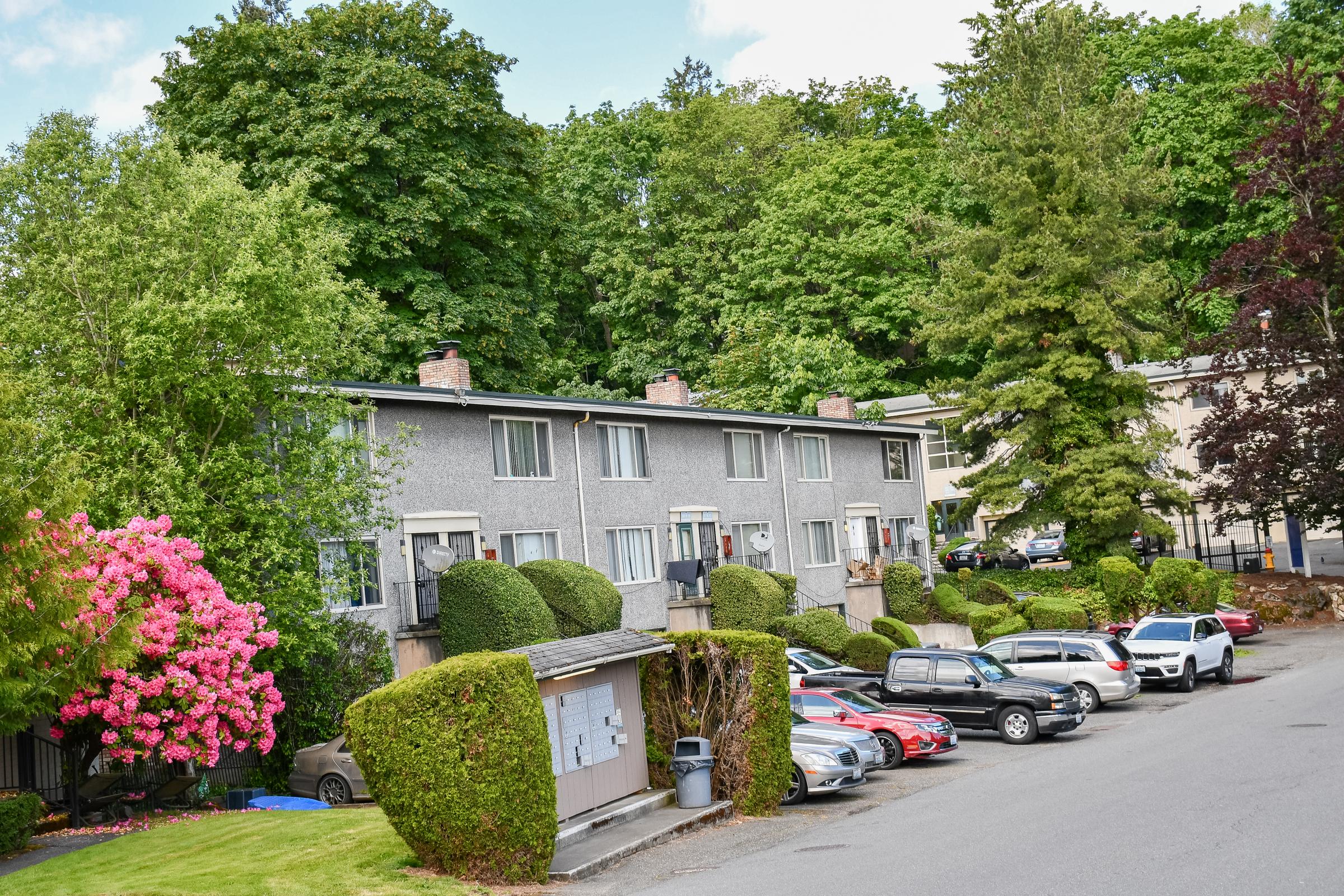A row of low-rise apartment buildings surrounded by lush greenery. In the foreground, several parked cars line a driveway. Vibrant pink flowering bushes and well-maintained hedges enhance the landscape, creating a welcoming atmosphere. The setting features a mix of trees and landscaped areas.