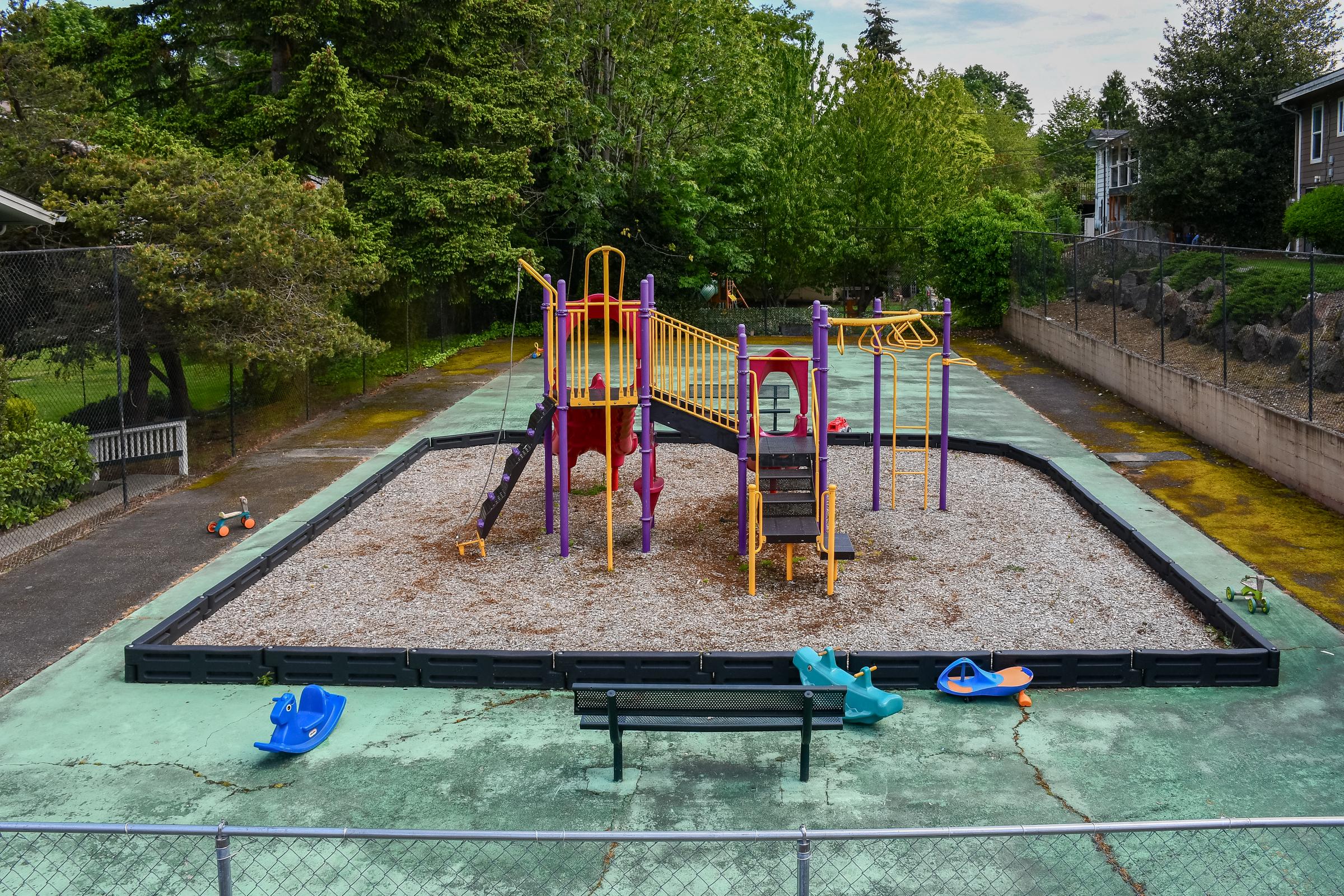 A colorful playground with slides, climbing structures, and swings situated on a gravel surface surrounded by green trees. The area features a bench in front and several small plastic toys scattered around. The playground is enclosed by a chain-link fence.