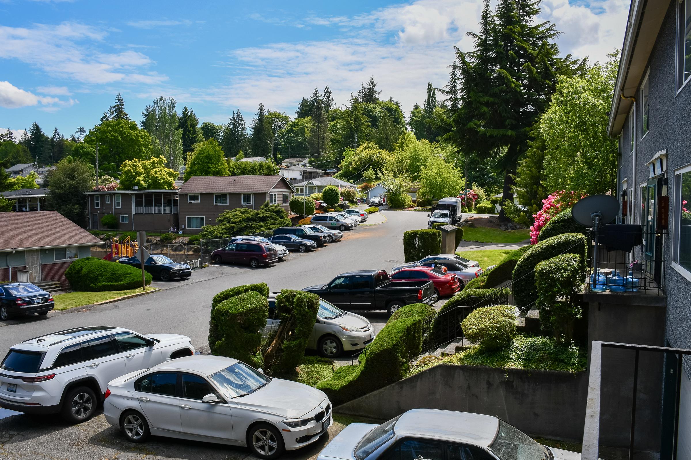View of a residential area with parked cars along the street. Lush green trees and shrubs frame the scene, while a few homes are visible in the background. The sky is partly cloudy, creating a bright and inviting atmosphere. The road curves gently, leading further into the neighborhood.