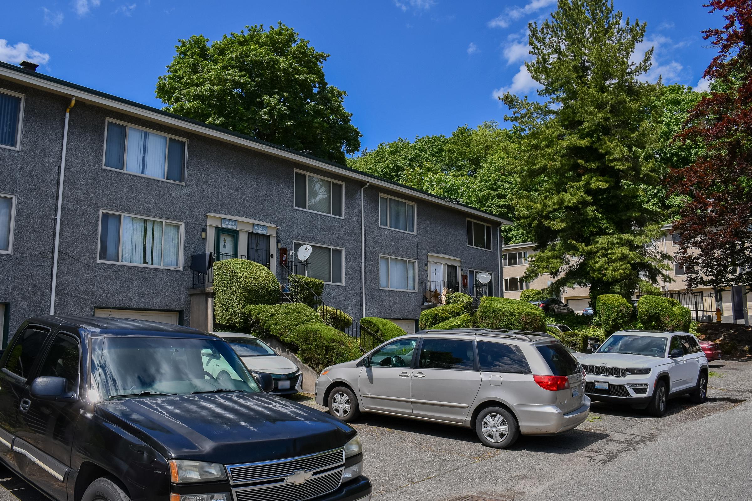 A residential apartment building with multiple units, featuring greenery and trees around it. Several parked cars, including a black truck, a silver minivan, and a white SUV, are lined up in front of the building under a clear blue sky with a few clouds.