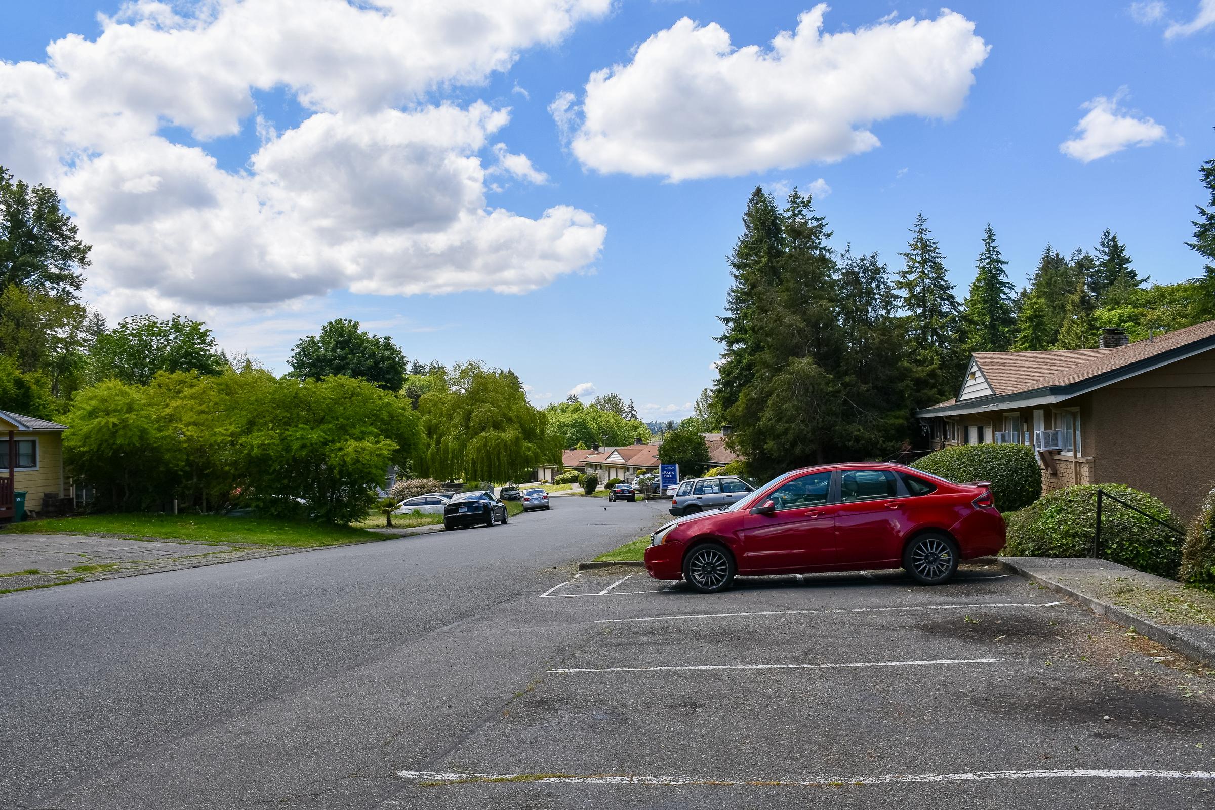 A quiet residential street with a few parked cars. A red car is visible in the foreground, while green trees and a row of houses line the street. Fluffy clouds float in a blue sky, creating a serene suburban atmosphere.