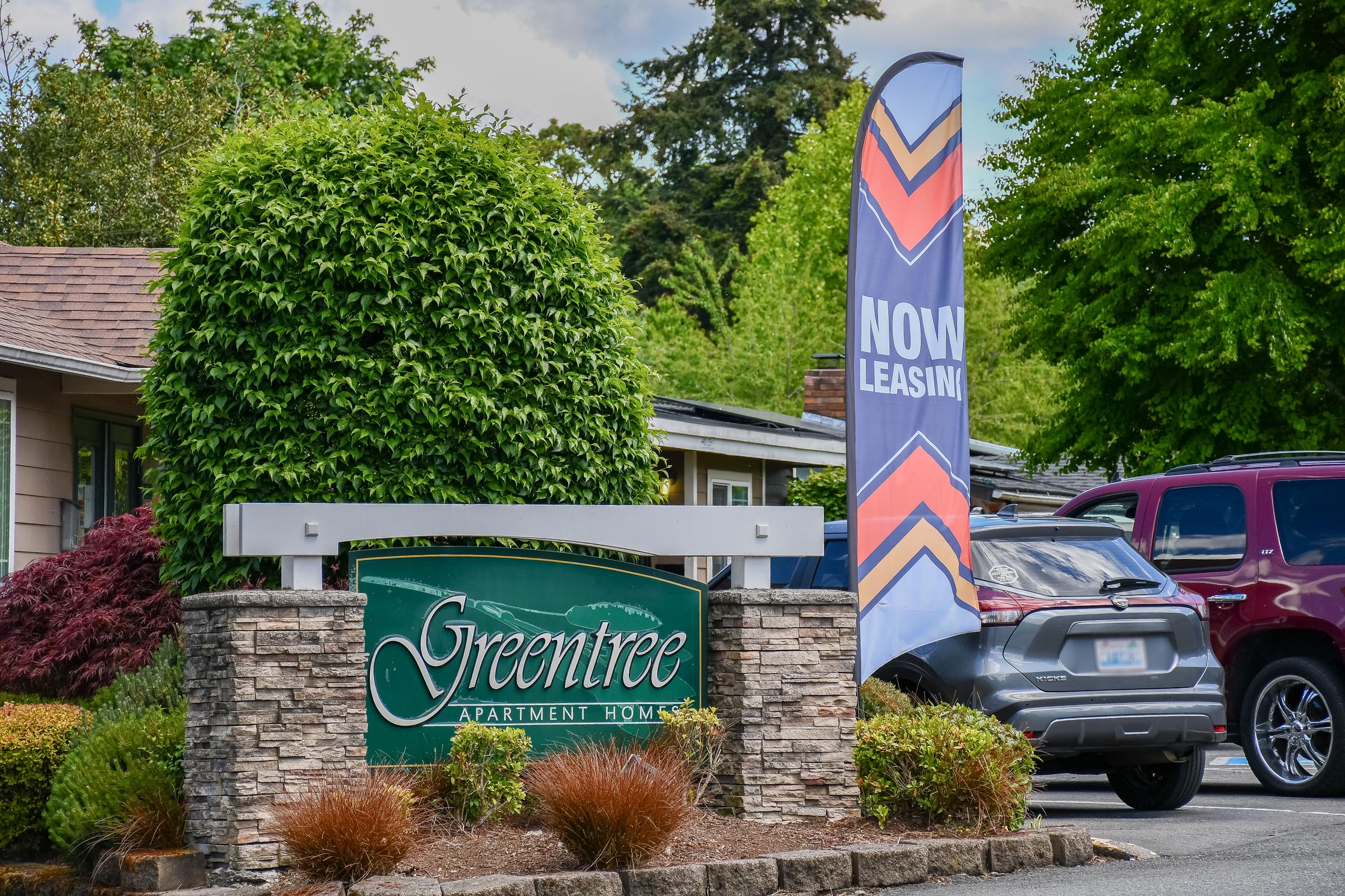Sign for Greentree Apartment Homes, featuring a green logo on a stone base. In the background, there are trees and shrubs, and a flag displaying "NOW LEASING" is visible. Two cars are parked nearby. The setting appears to be an apartment complex with well-maintained landscaping.