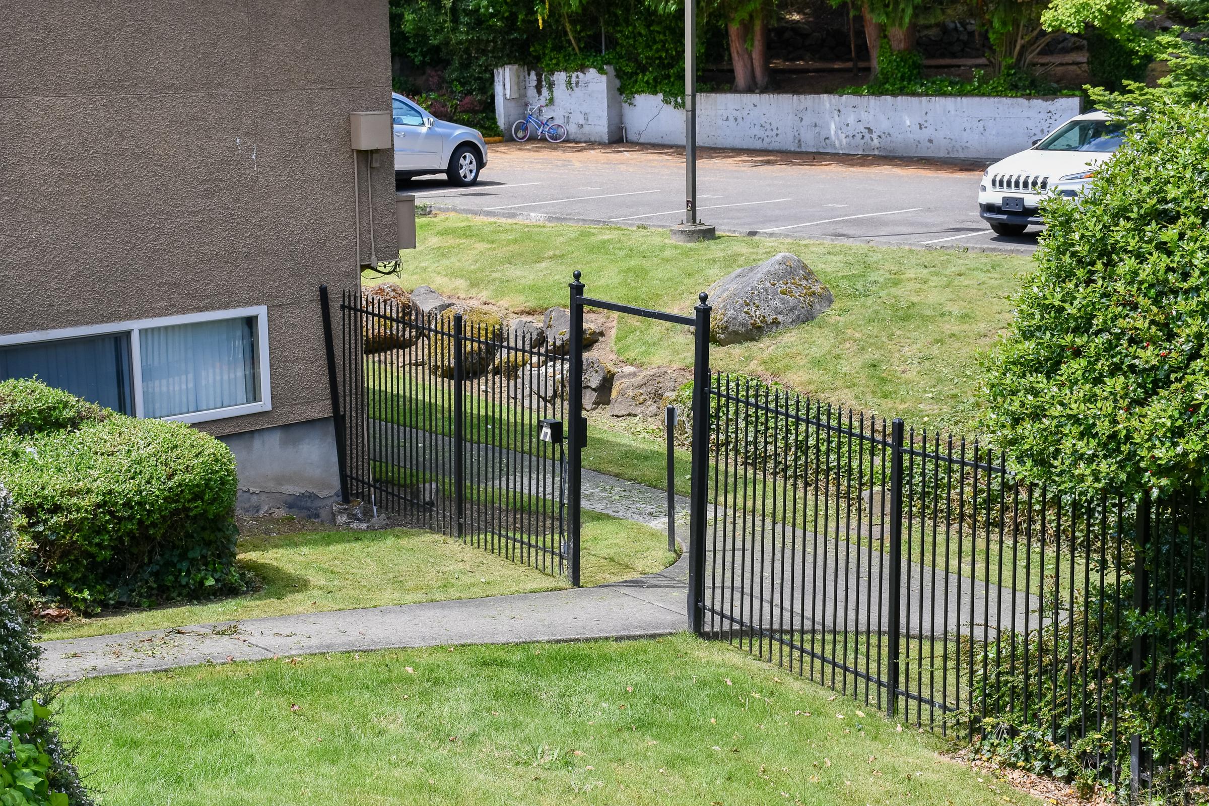A view of a gated entrance to a pathway, surrounded by grassy areas and bushes. In the background, a parking lot is visible with some vehicles, and trees provide greenery. The scene is well-lit and conveys a peaceful outdoor setting.