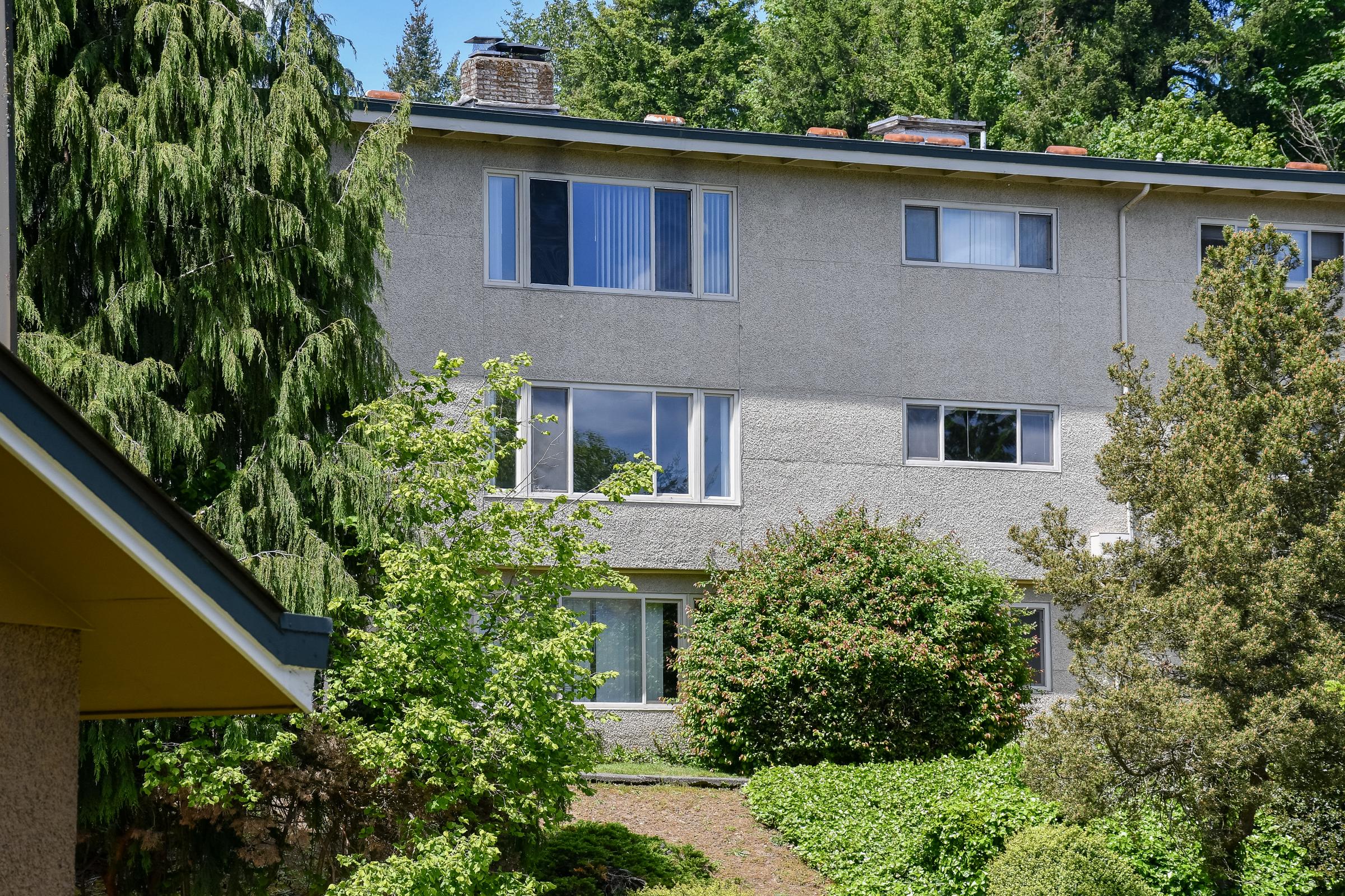 A two-story building with a textured gray exterior surrounded by greenery. The left side of the image shows a low, slanted roof and branches from trees. Windows are visible on the front of the building, with some partially obscured by leaves, creating a serene, natural setting.