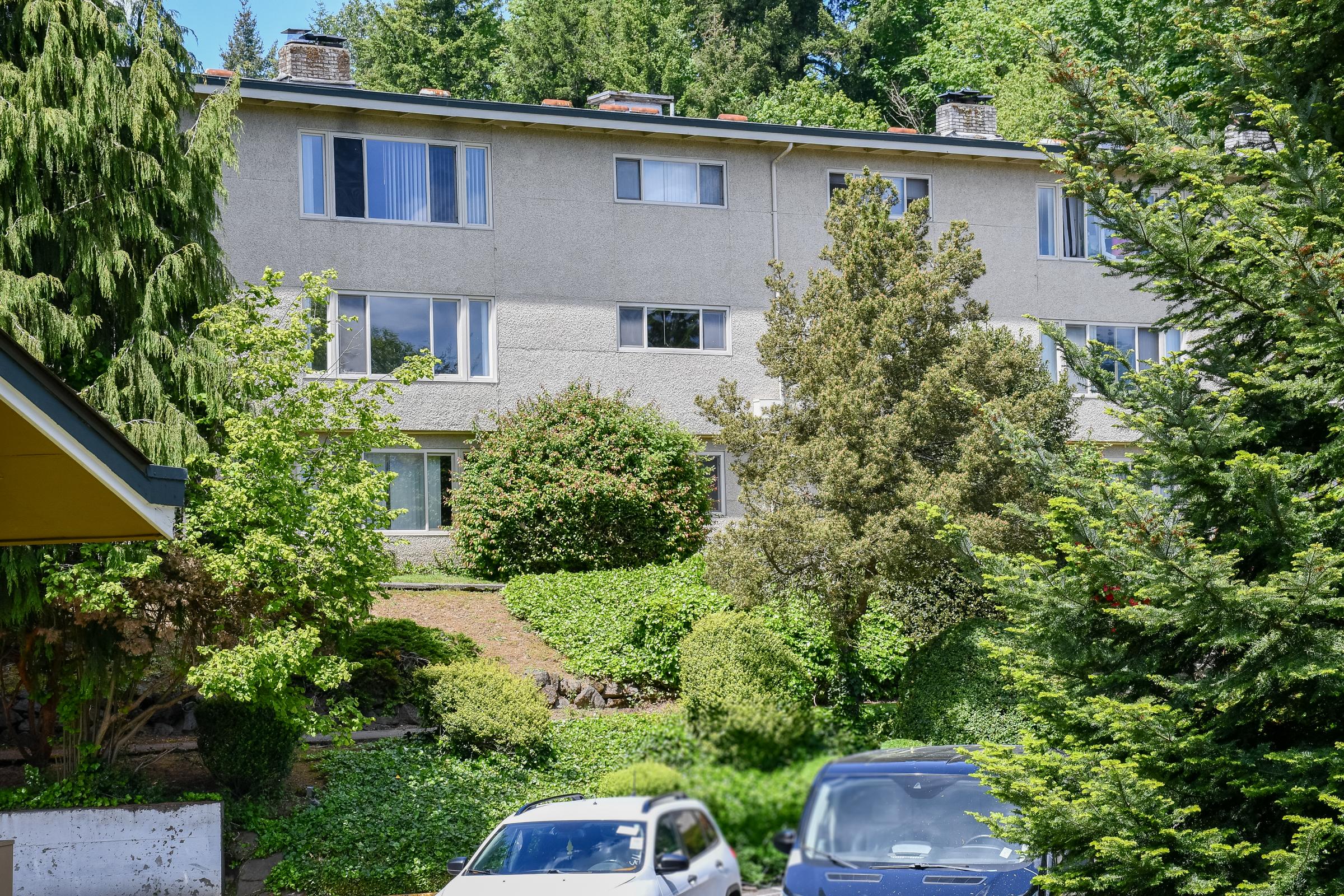 A multi-story residential building surrounded by greenery, including trees and bushes. In the foreground, two vehicles are parked on a driveway, with a well-maintained garden area visible. The building has several windows reflecting sunlight.