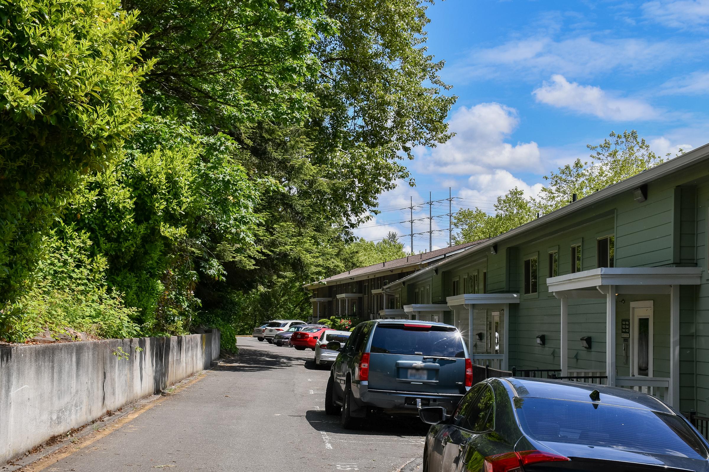 A quiet residential street lined with green trees and bushes, featuring multiple parked cars along the side. On one side, there are modern two-story apartment buildings with light green exteriors under a clear blue sky dotted with clouds.