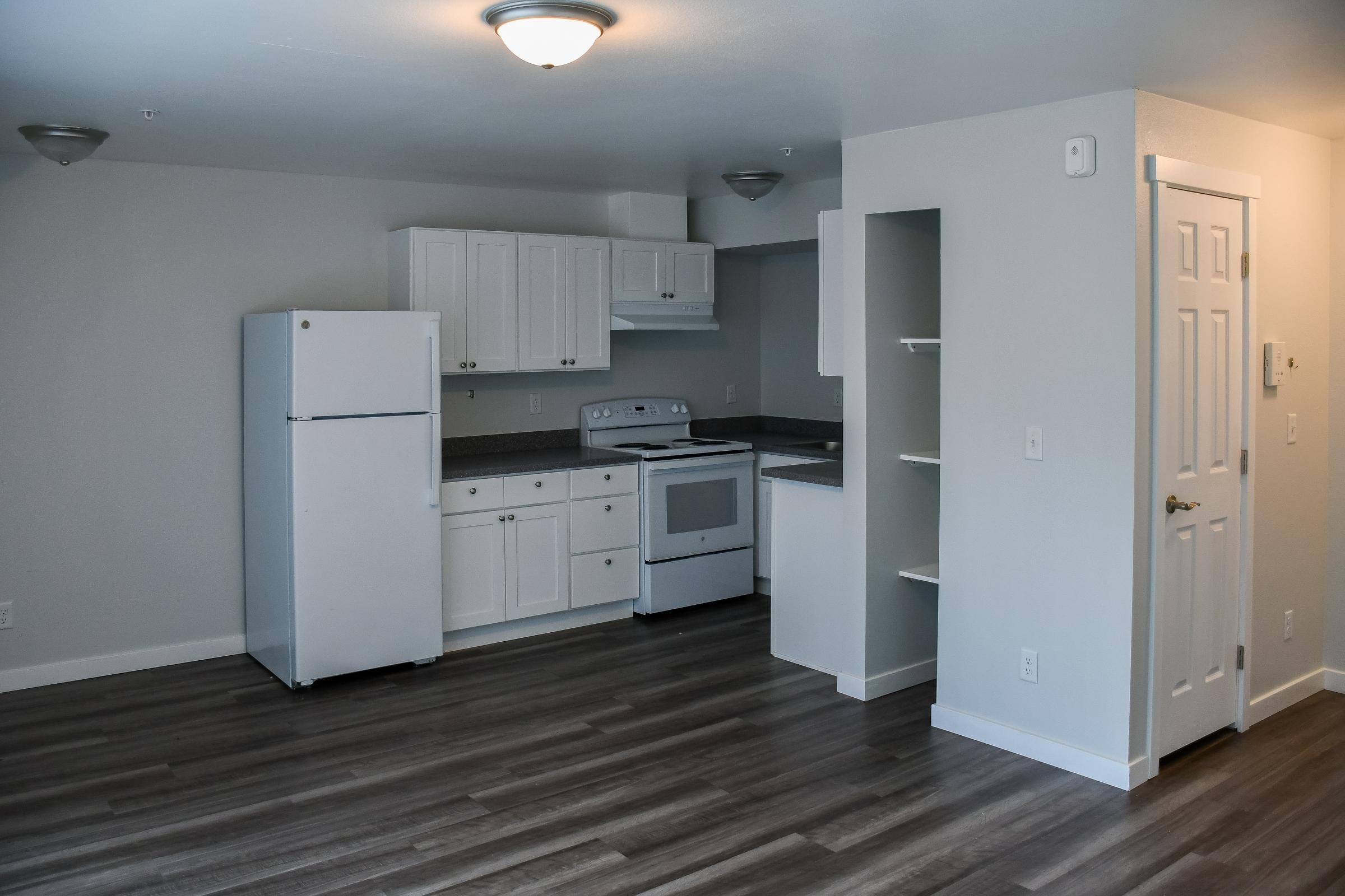 A modern kitchen and living area featuring white cabinetry, a refrigerator, an oven, and a countertop. The space has gray walls, wood-like flooring, and bright overhead lighting. A door leads to another room, and there’s a built-in shelf in the corner, creating an open and inviting atmosphere.