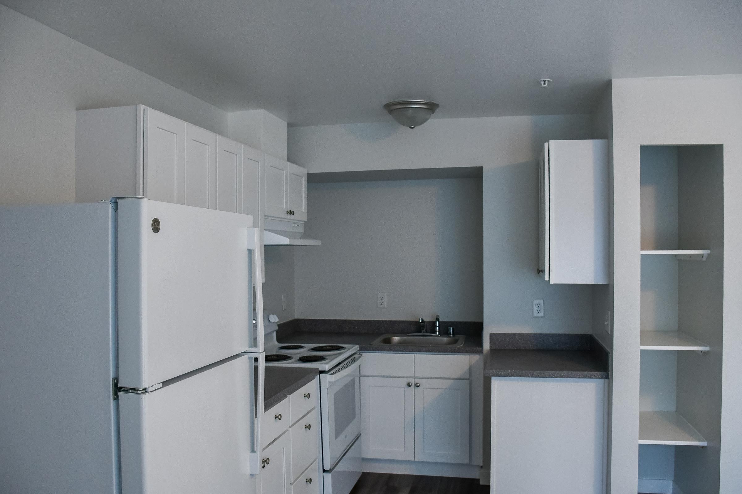 A modern kitchen featuring white cabinetry, a refrigerator, an oven, and a sink. The countertops are gray, and there are shelves for storage. The design is minimalist, with neutral wall colors and a clean, spacious layout.