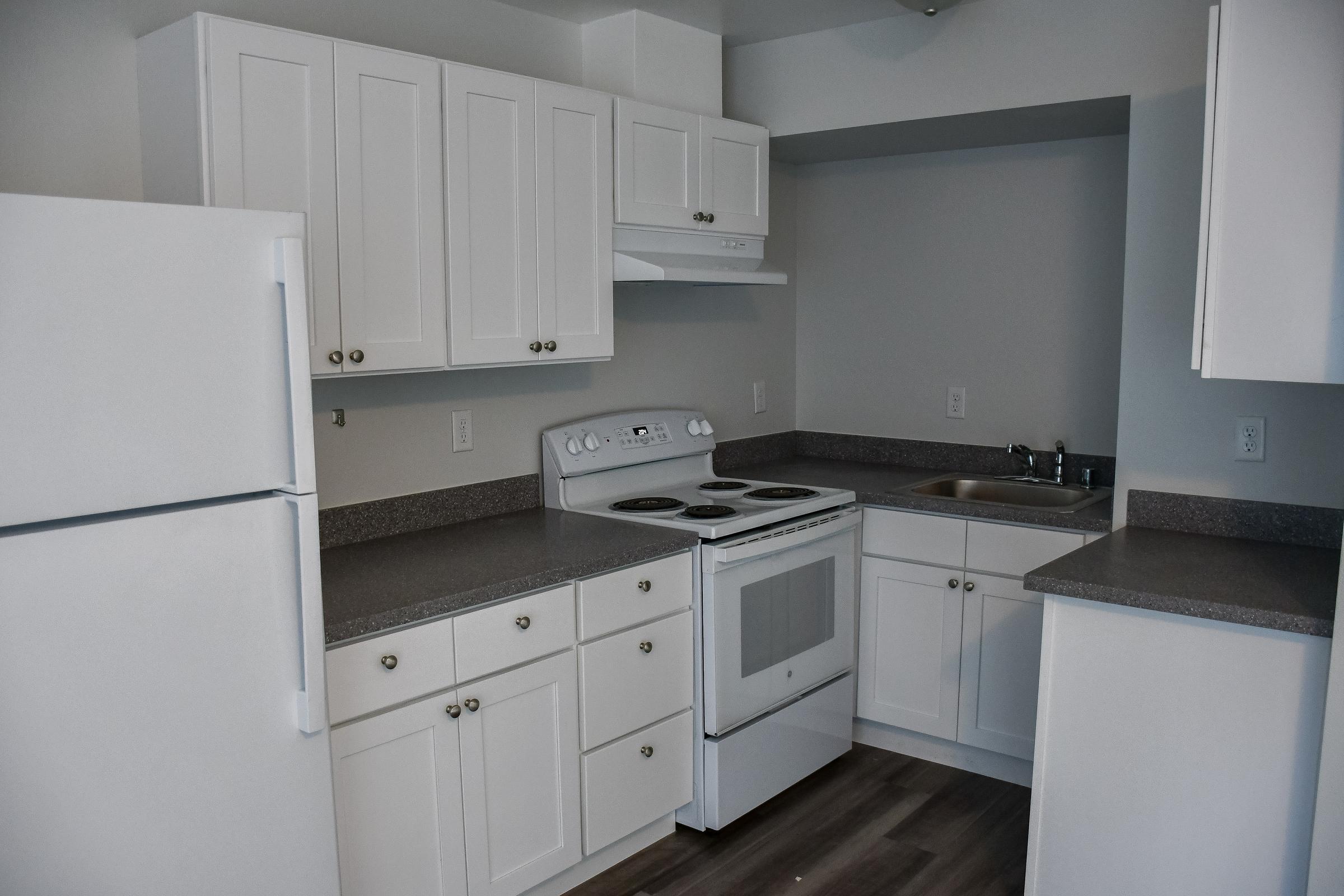 A modern kitchen featuring white cabinetry, a stovetop and oven, a sink, and a refrigerator. The countertops are gray, and the walls are a light color. The space is well-lit and clean, with a minimalist design that conveys a contemporary look.