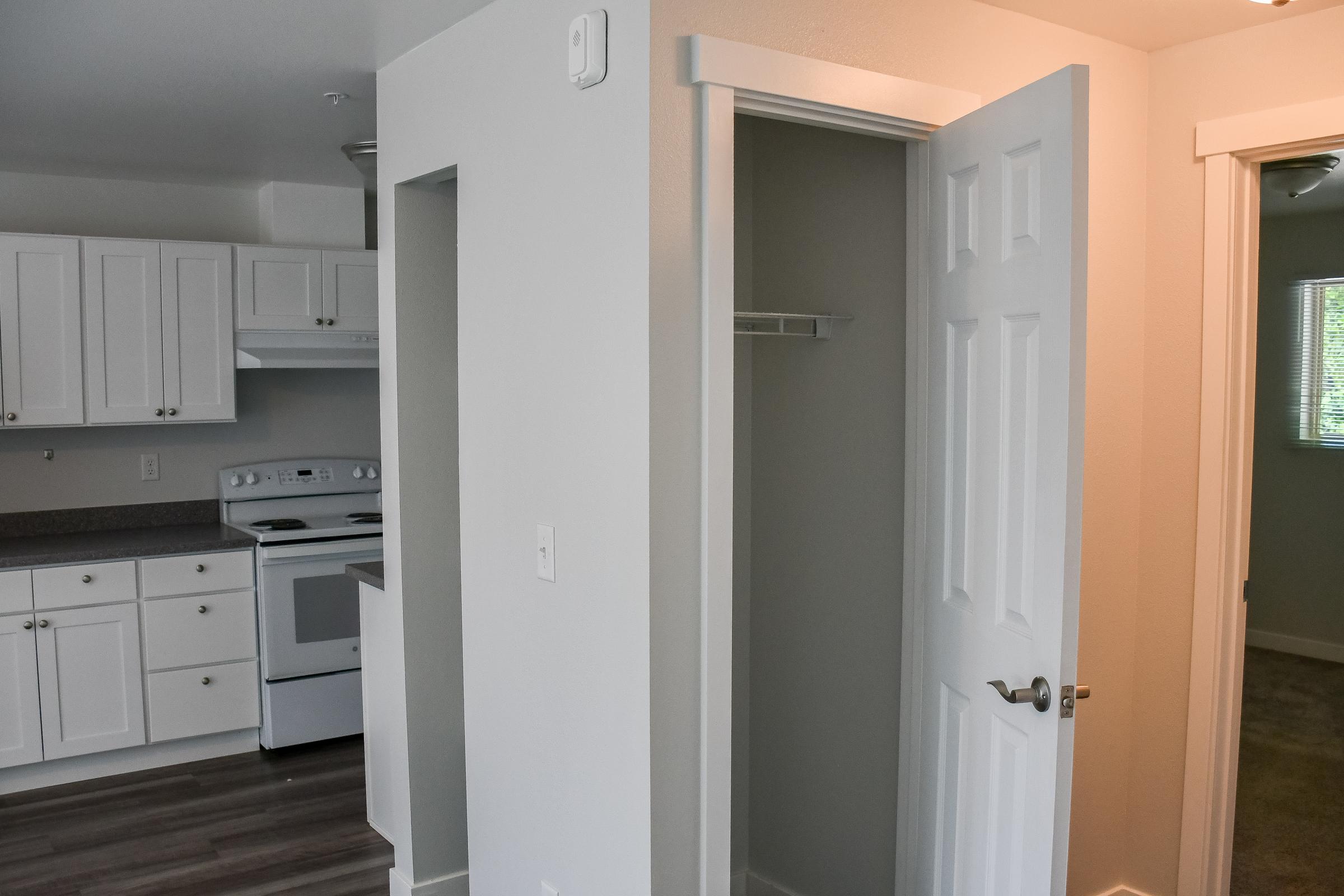 A view of a modern kitchen featuring white cabinets, a stove, and counter space. In the foreground, there is an open door leading to a small closet. The walls are painted in light colors, creating a bright and airy atmosphere, with a glimpse of another room visible in the background.