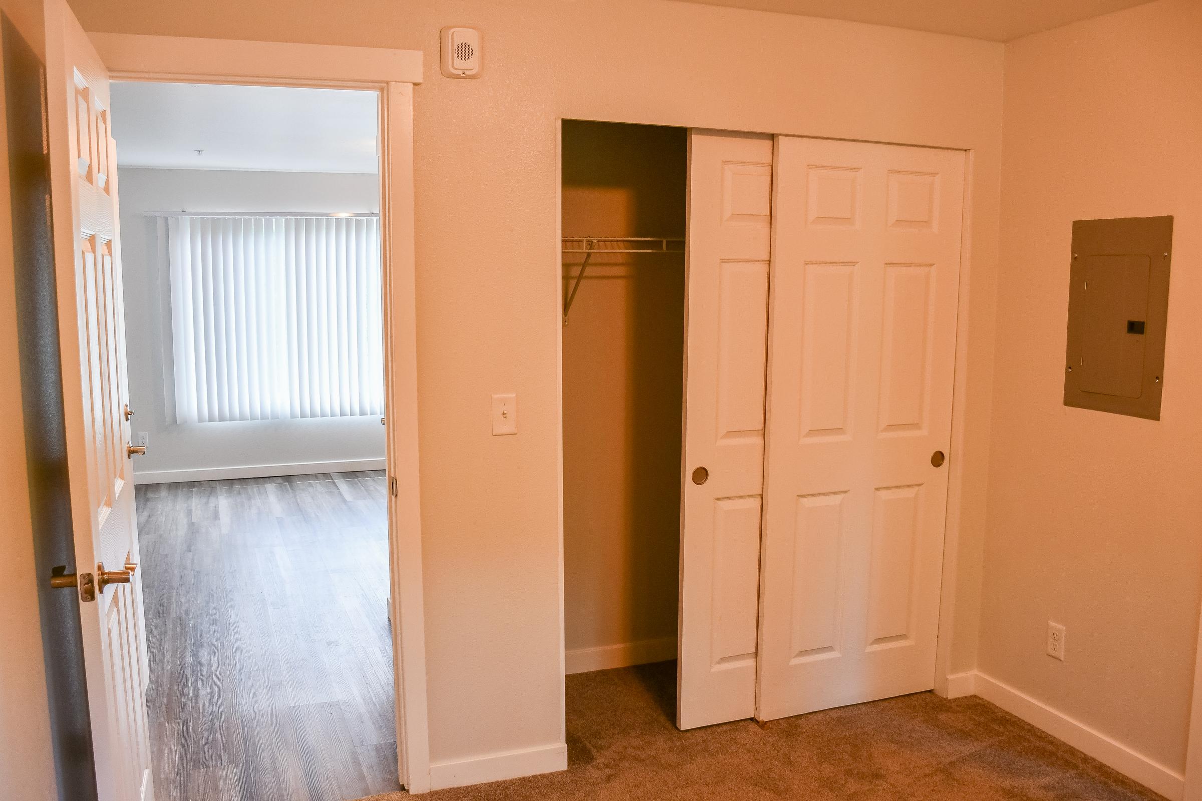 An interior view of a room featuring two doors: one leading to another space with window coverings and the other opening to a closet with a shelf. The flooring is a mix of carpet and wood, and the walls are painted a neutral color.