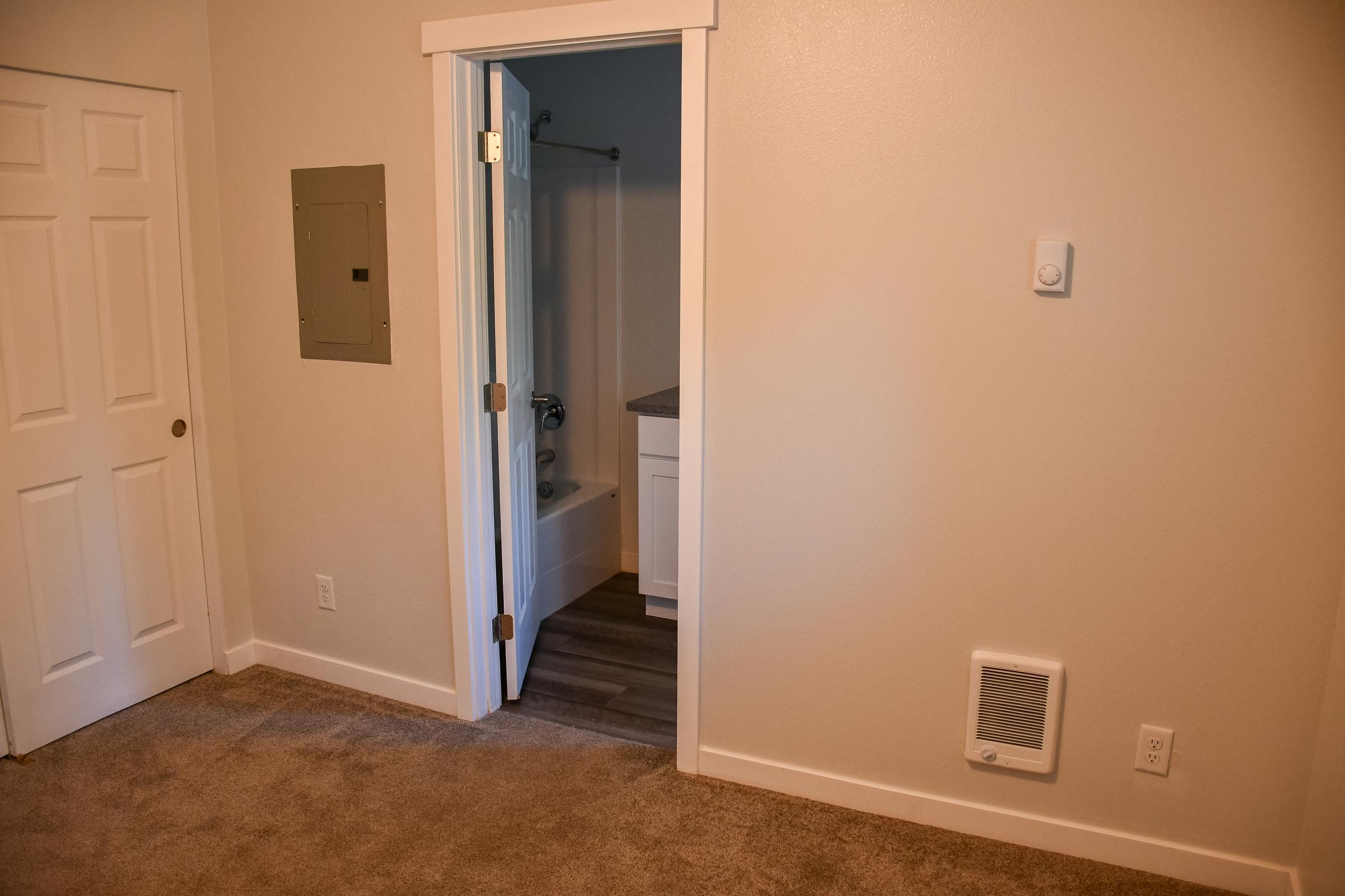 A simple room featuring beige walls and carpet. On the left, there’s a closed white door, while an open doorway leads to a bathroom with a bathtub visible inside. To the right, a wall-mounted heating vent can be seen, and there's a small utility panel on the wall. Natural light illuminates the space.