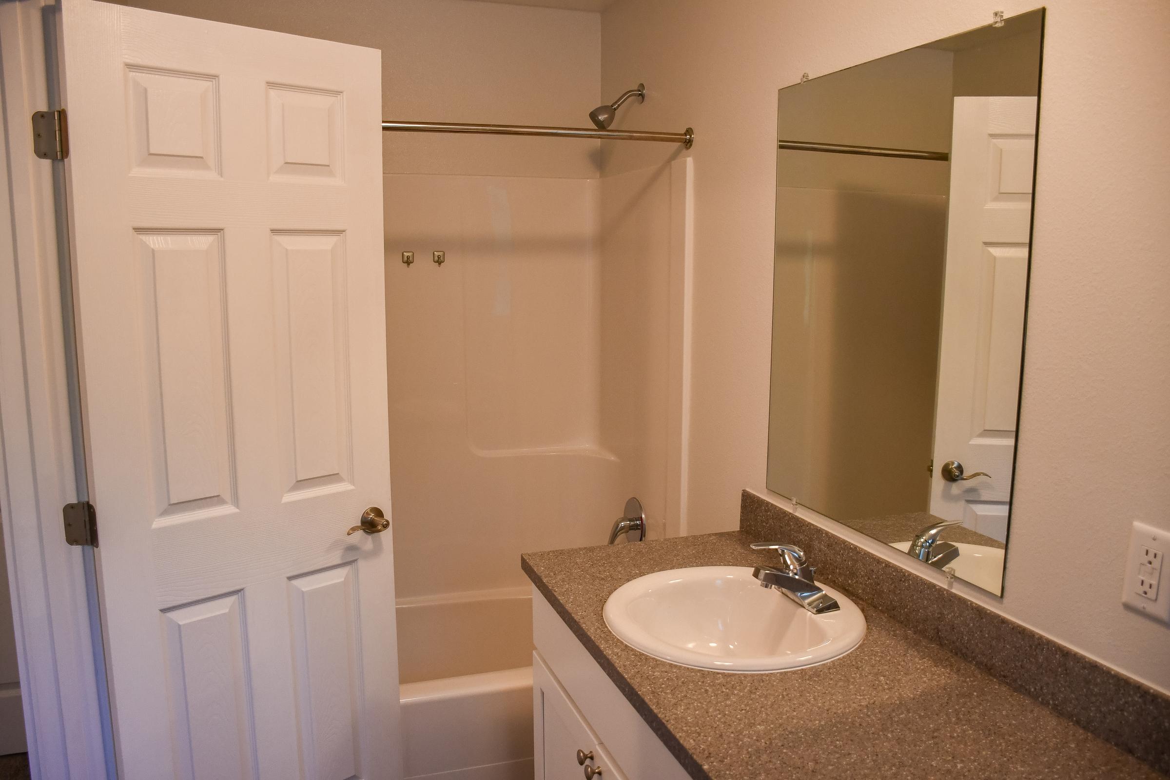 A bathroom featuring a shower tub combo, a single sink vanity with a faucet, and a mirror above the sink. The walls are painted light colors, and there is a white door partially opened, providing access to the bathroom. The flooring appears to be tiled.