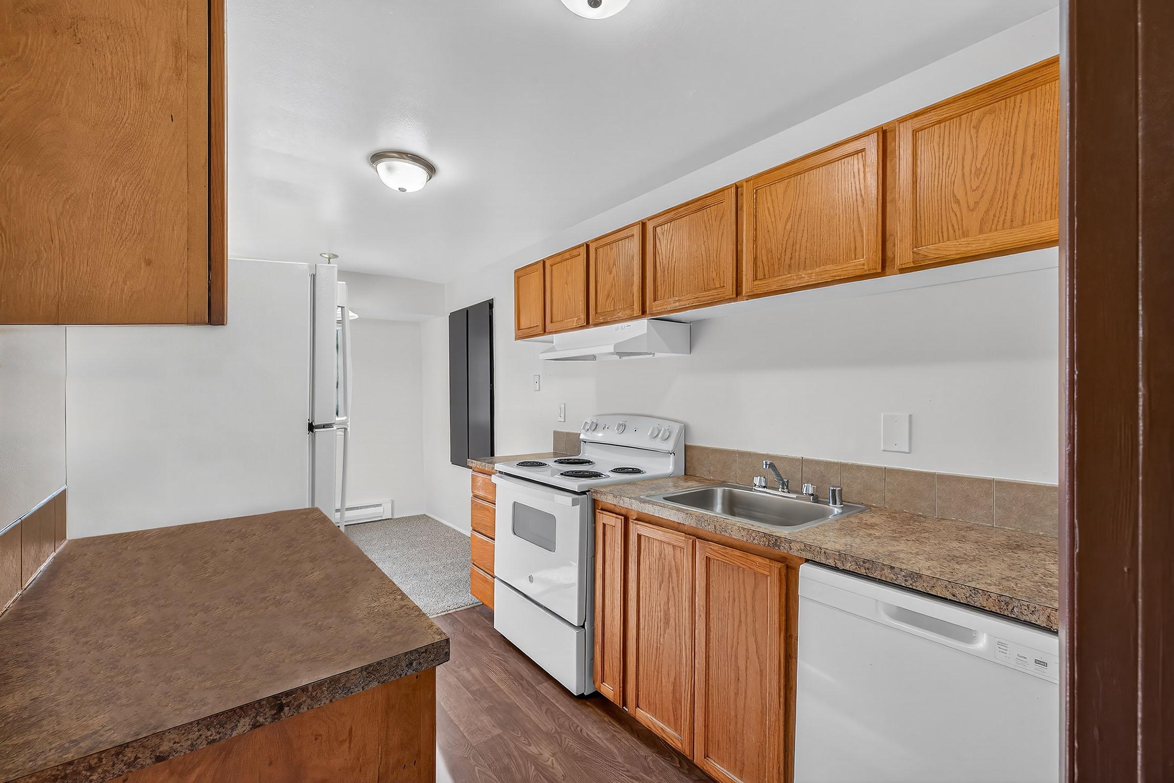A modern kitchen featuring wooden cabinetry, a white stove, a sink, and a dishwasher. The countertops are brown, and the flooring is a dark wood finish. The space is well-lit with overhead lighting, and there's a glimpse of a nearby room in the background.