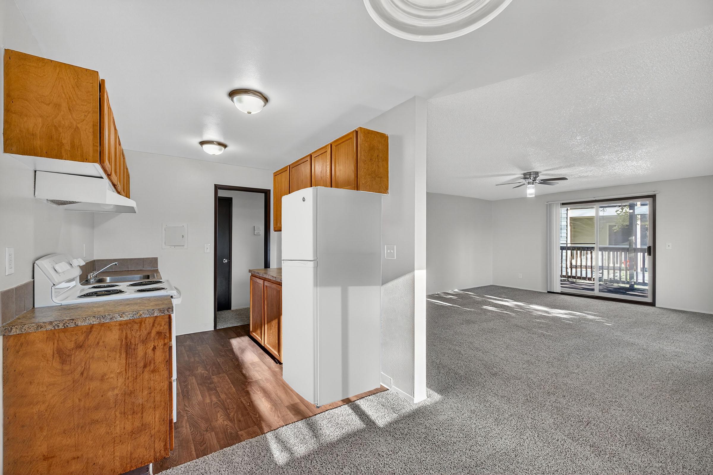 A view of a cozy apartment interior featuring a kitchen with brown wooden cabinets, a white refrigerator, and a small stove. Adjacent is a spacious living area with light-colored walls and a ceiling fan, alongside a balcony door leading to an outdoor space. Soft carpets cover the floor.