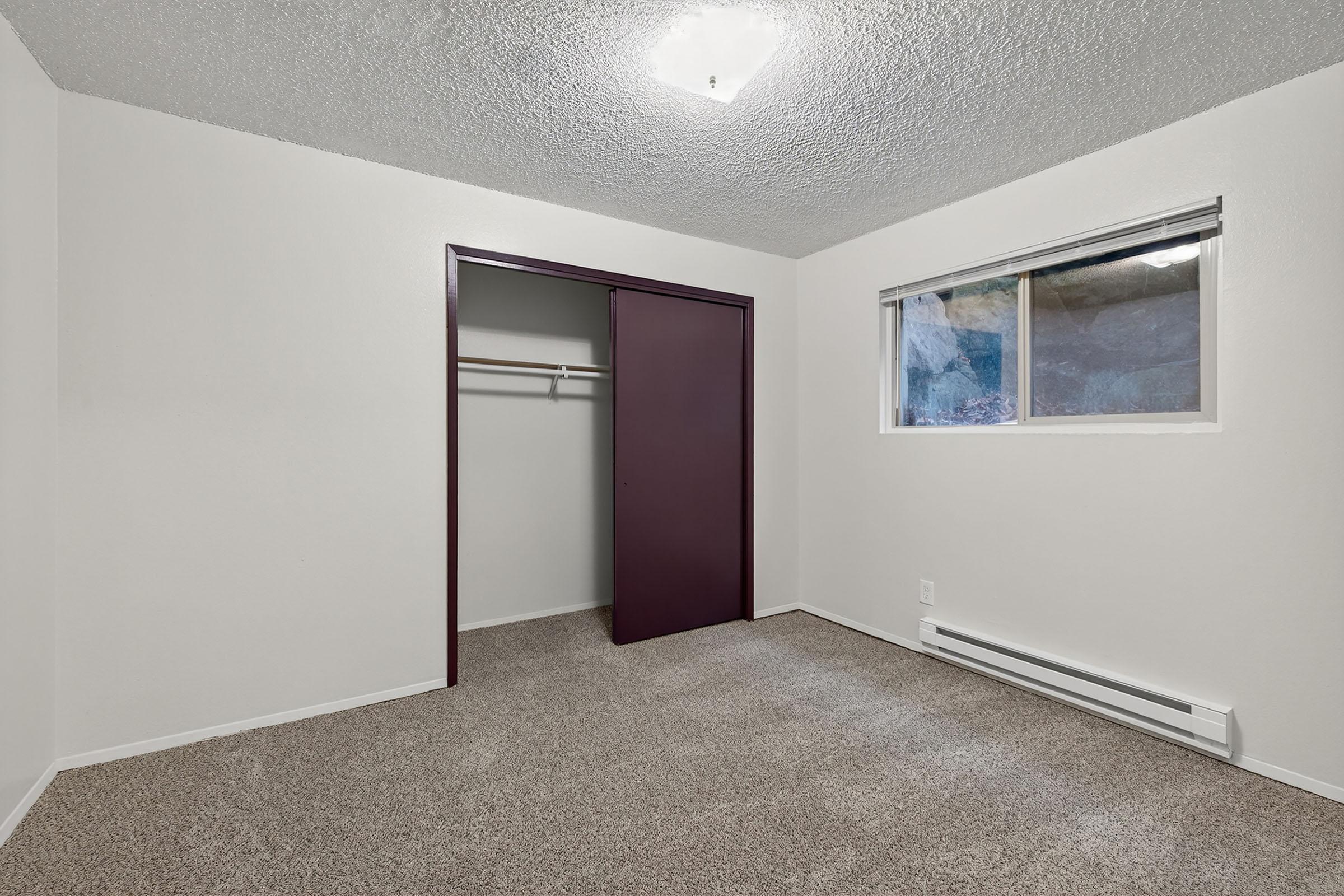 A clean, empty bedroom featuring light-colored walls, a textured ceiling, and carpeted flooring. There is a window letting in natural light, and a closet with a closed door on one side. The space appears well-maintained and ready for new furnishings.