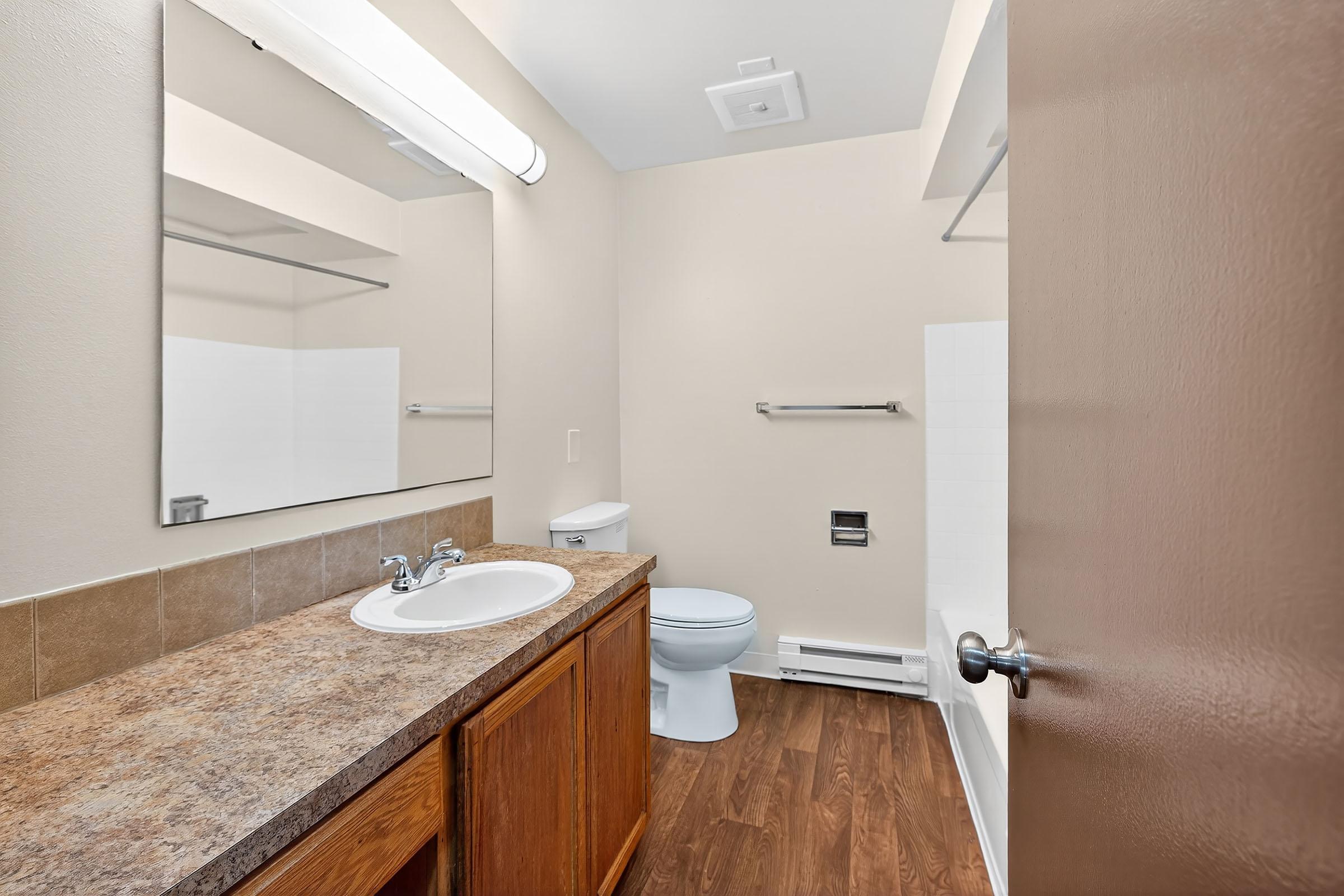 A clean, modern bathroom featuring a white toilet, a sink with a countertop, and a large mirror. Warm wood cabinetry complements the neutral-colored walls and flooring. A bath area with a white shower and tub is visible, with a towel rack and lighting above the mirror.
