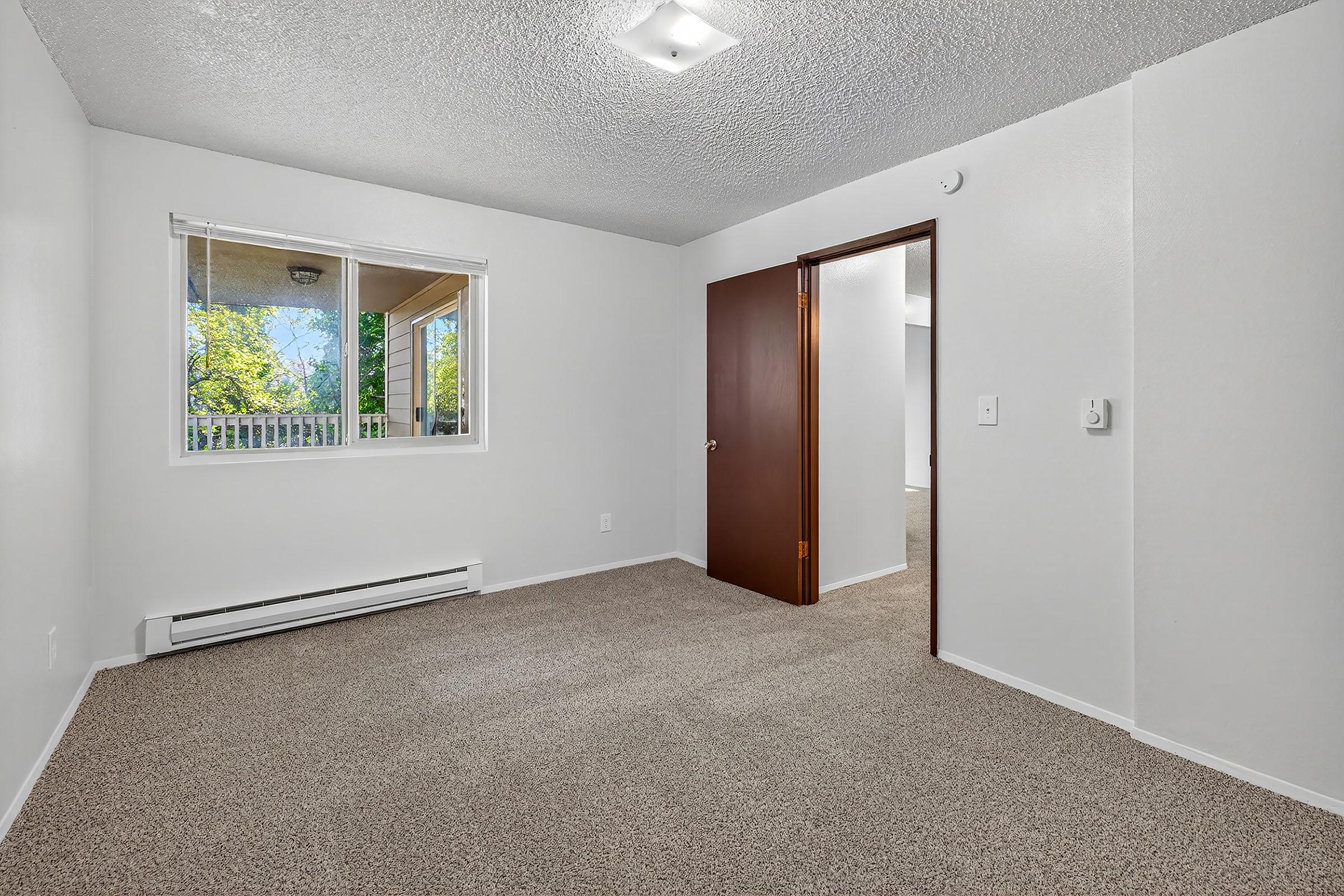 A vacant room with light-colored walls and beige carpet. It features a window letting in natural light, a door leading to another room, and a heating unit on the wall. The ceiling has a simple overhead light fixture, creating an airy and spacious feel.